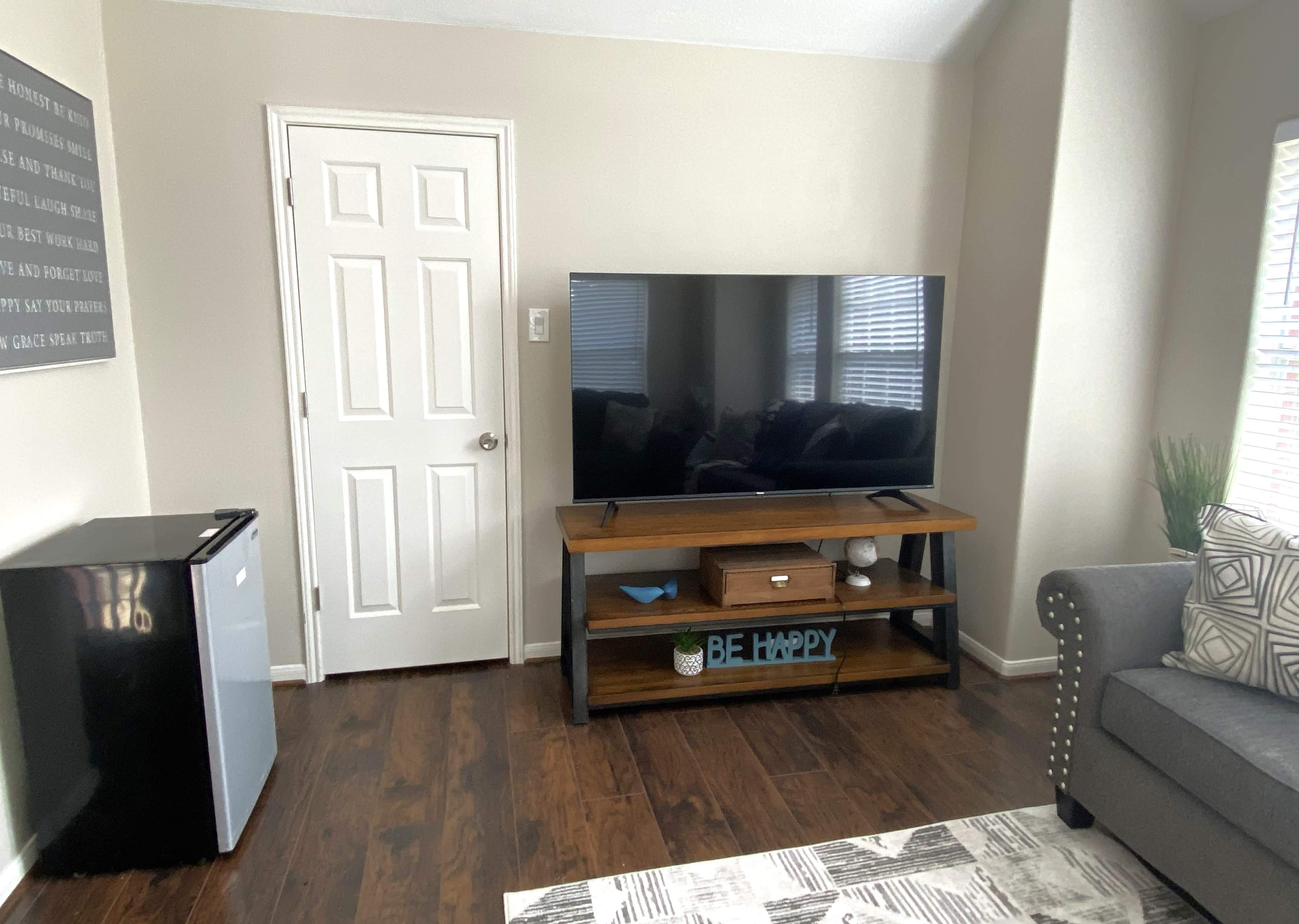A living area with a large TV on a wooden stand, a small refrigerator, a door, and a comfortable armchair.
