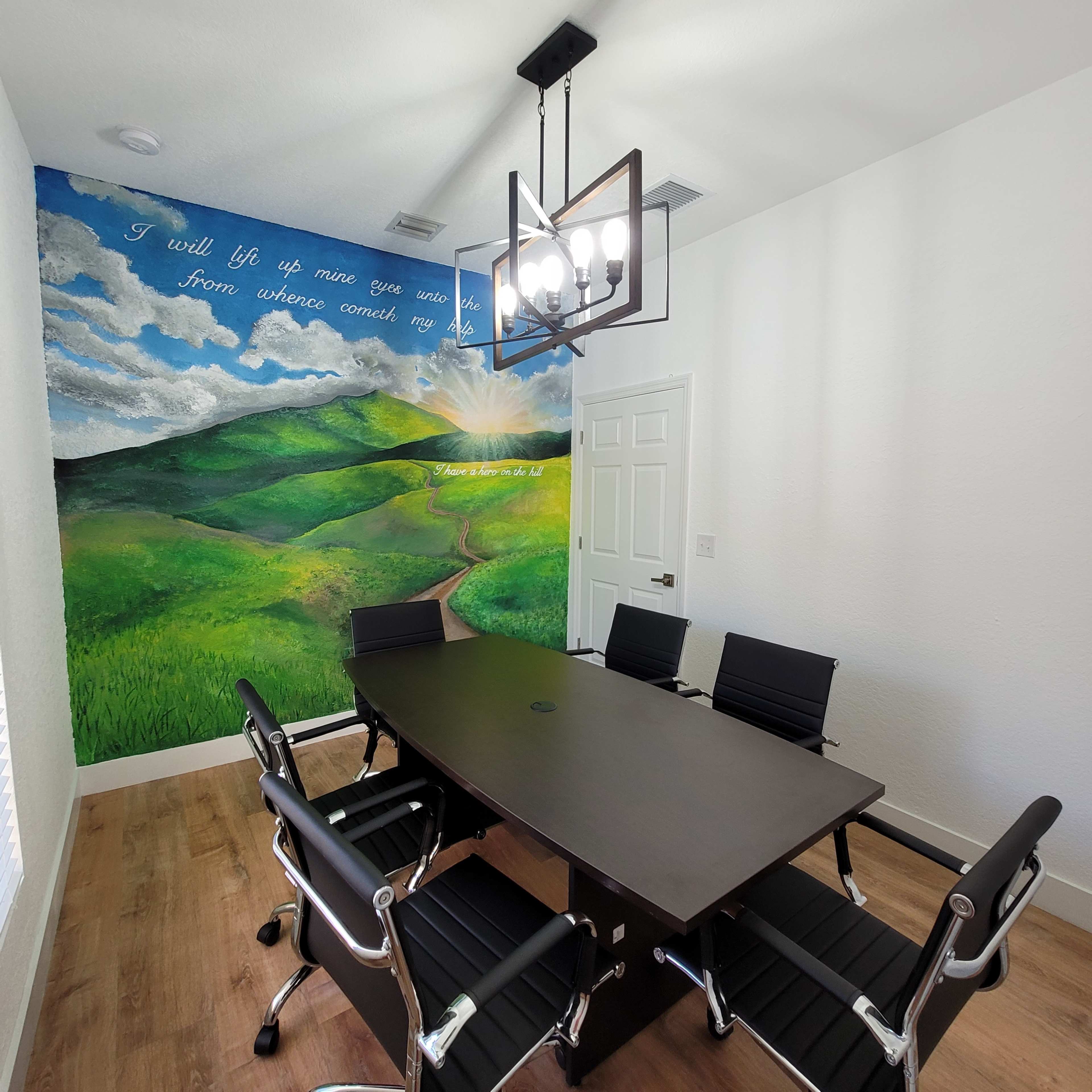 A conference room with a large table surrounded by black chairs and a mural of green hills on one wall featuring an inspirational quote.