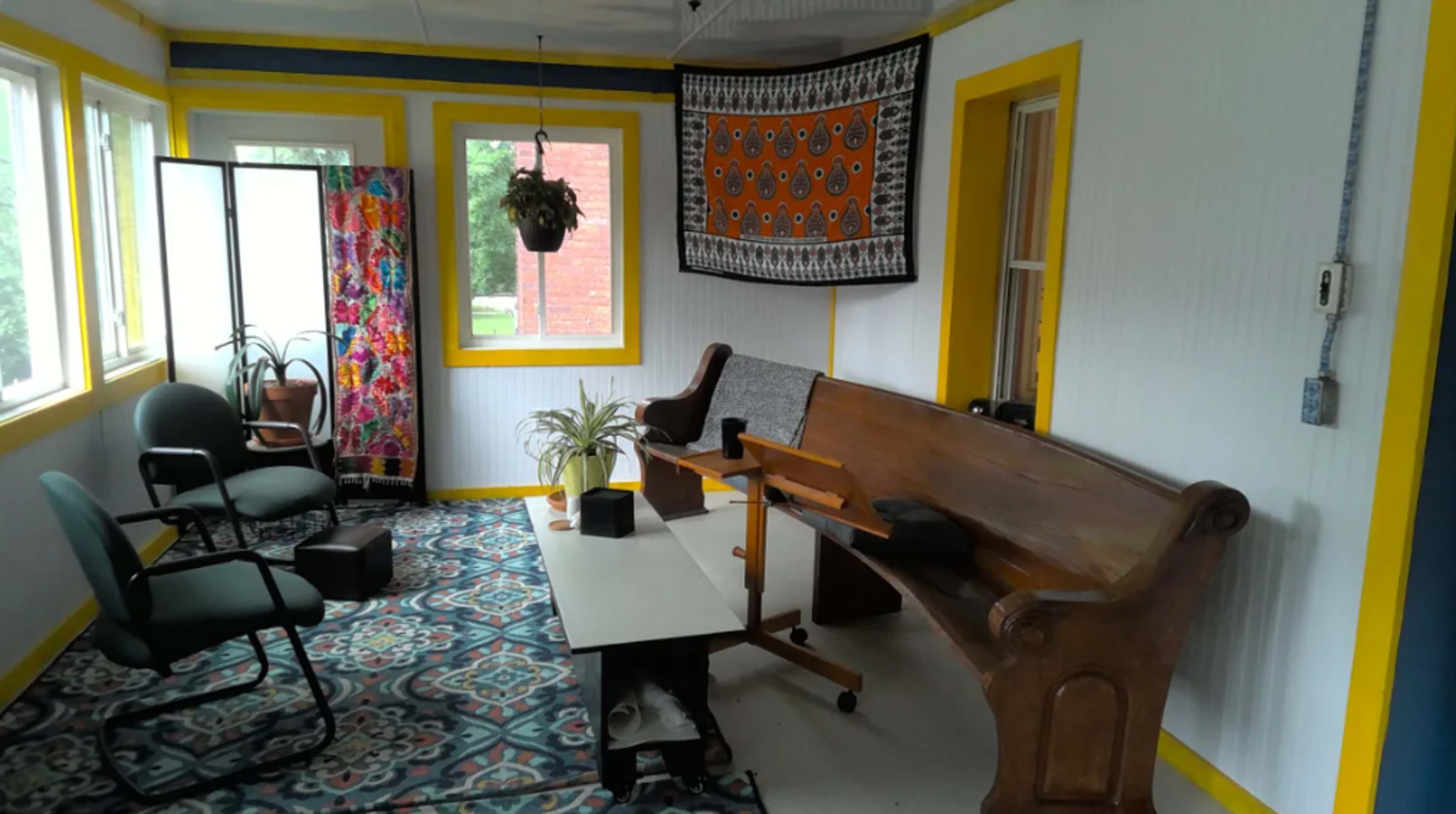 A sunlit room with colorful decor, featuring two chairs, a bench, a table, and a patterned rug.