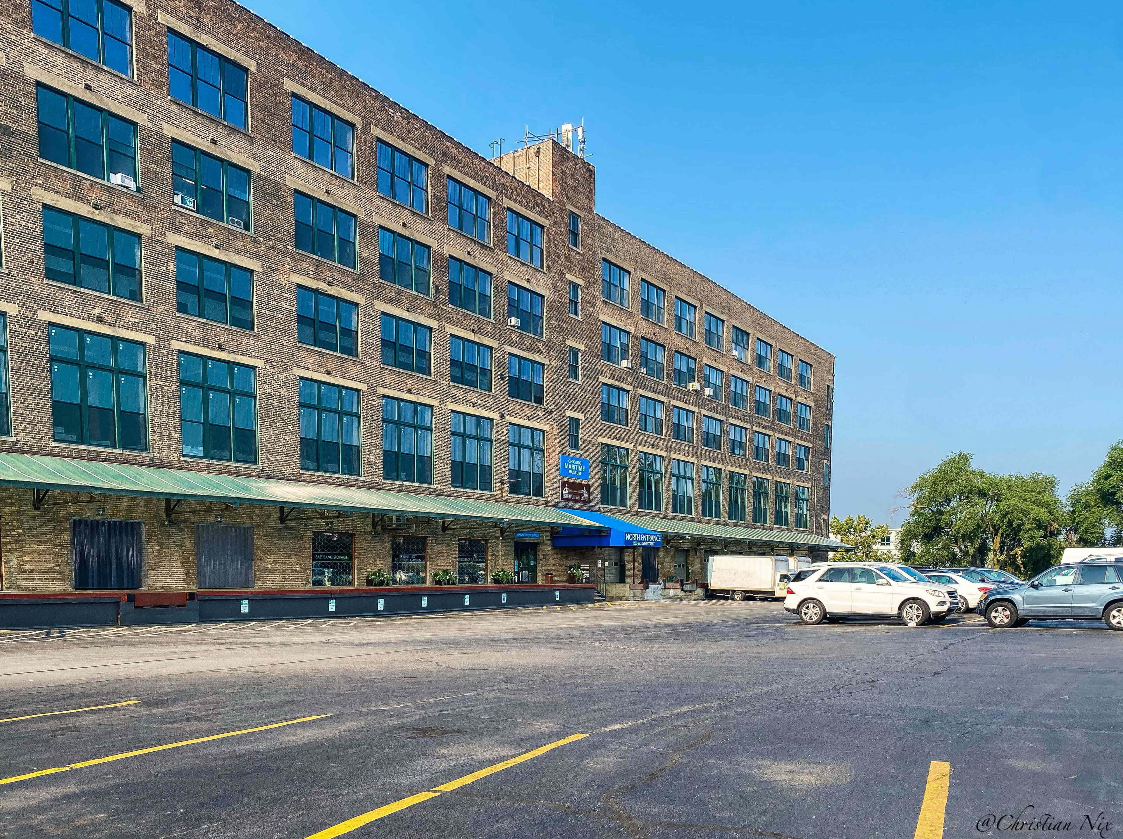 The image shows a multi-story brick building with large windows and a loading area, accompanied by parked vehicles in front.