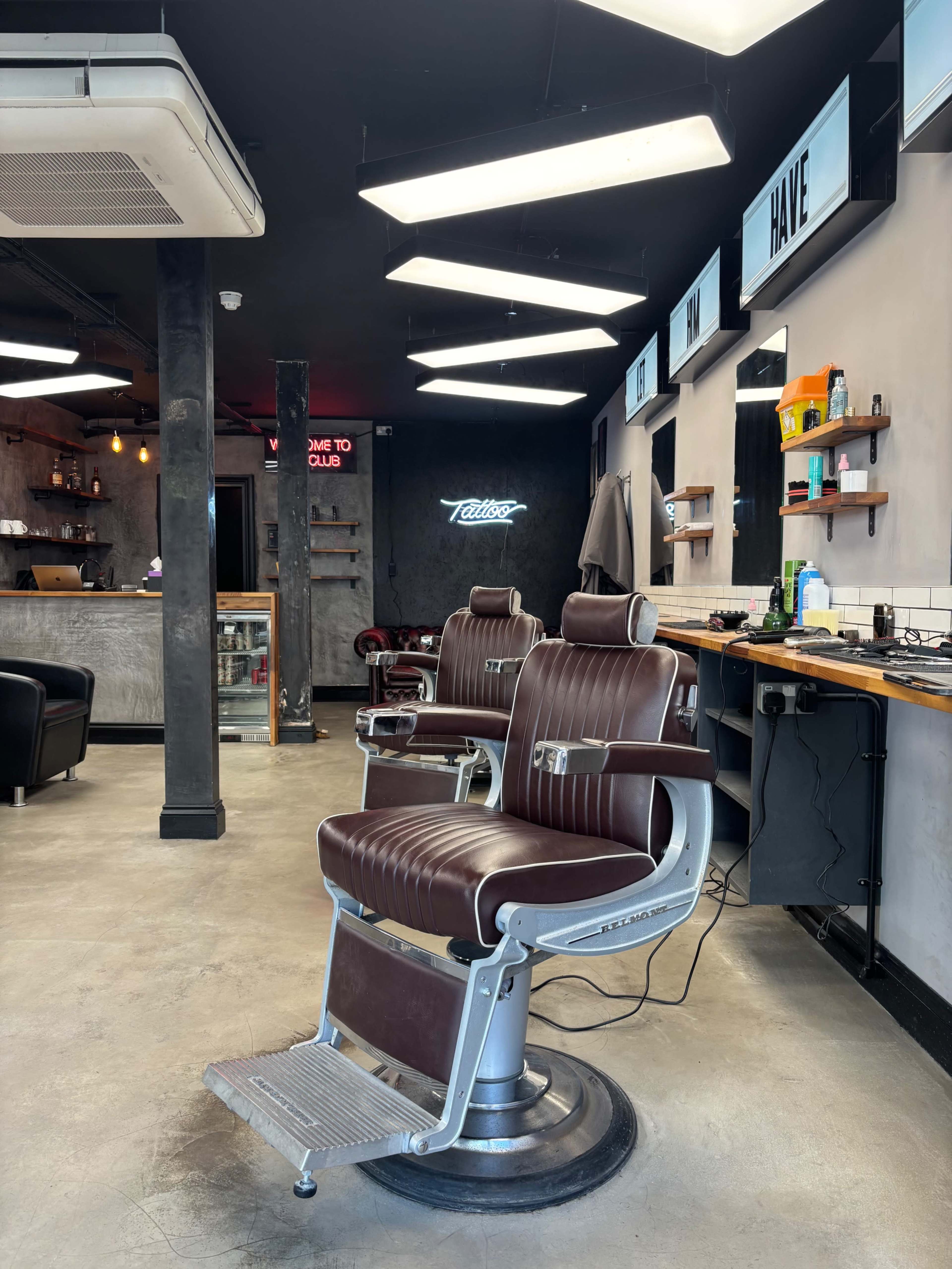 The image shows a modern barbershop interior with two barber chairs, a counter with grooming products, and a neon sign in the background.