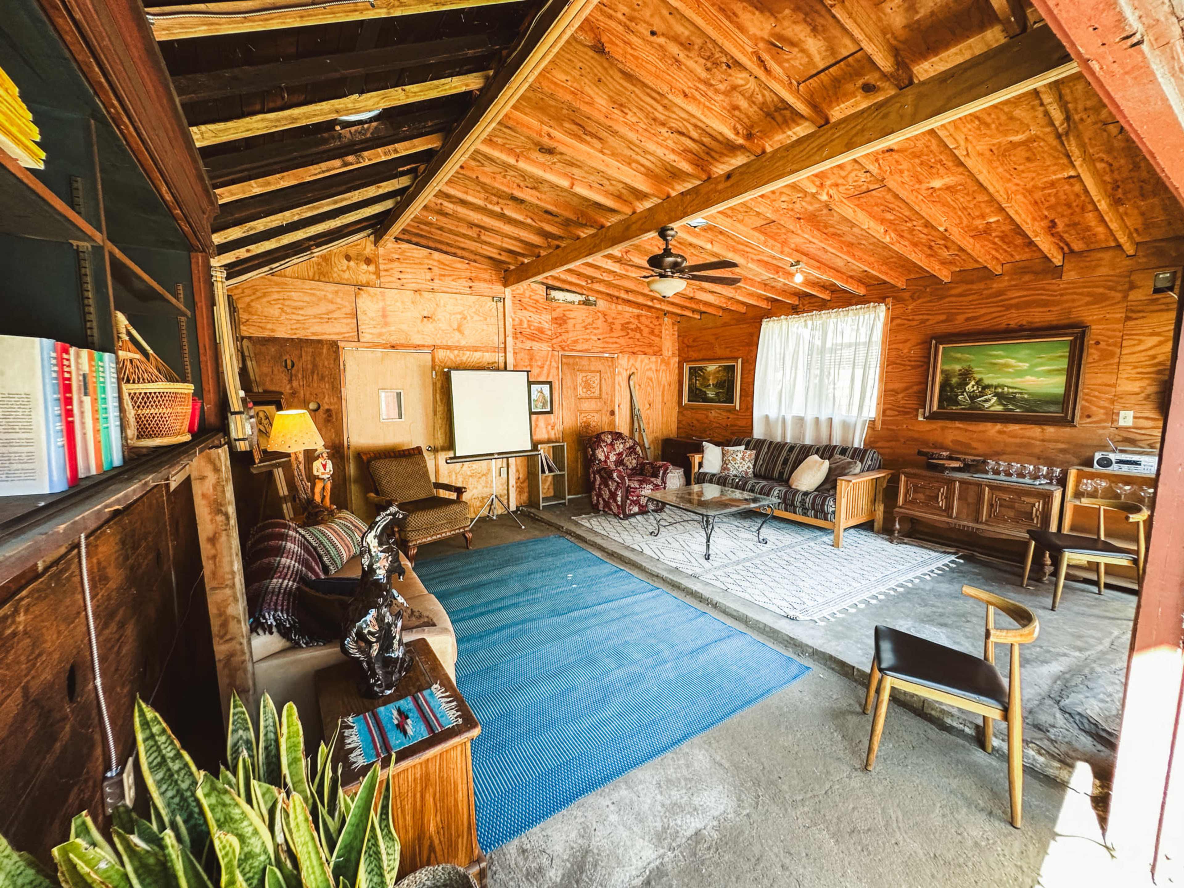 The interior of a rustic cabin features wooden walls, a ceiling fan, a seating area with various furniture, and a large window letting in natural light.