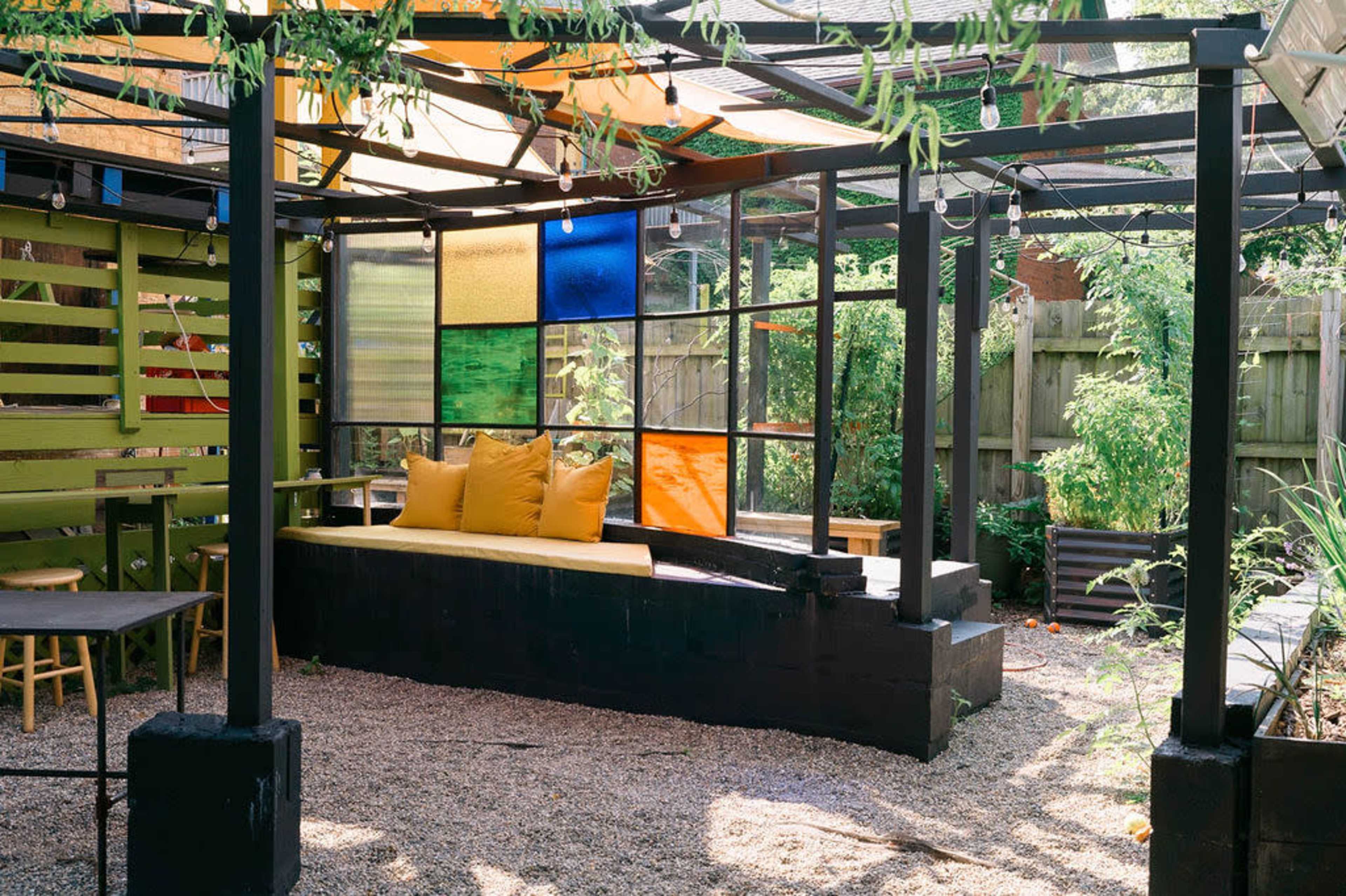 A modern outdoor seating area features a large, colorful glass panel wall, a yellow cushioned bench, and gravel flooring surrounded by greenery.