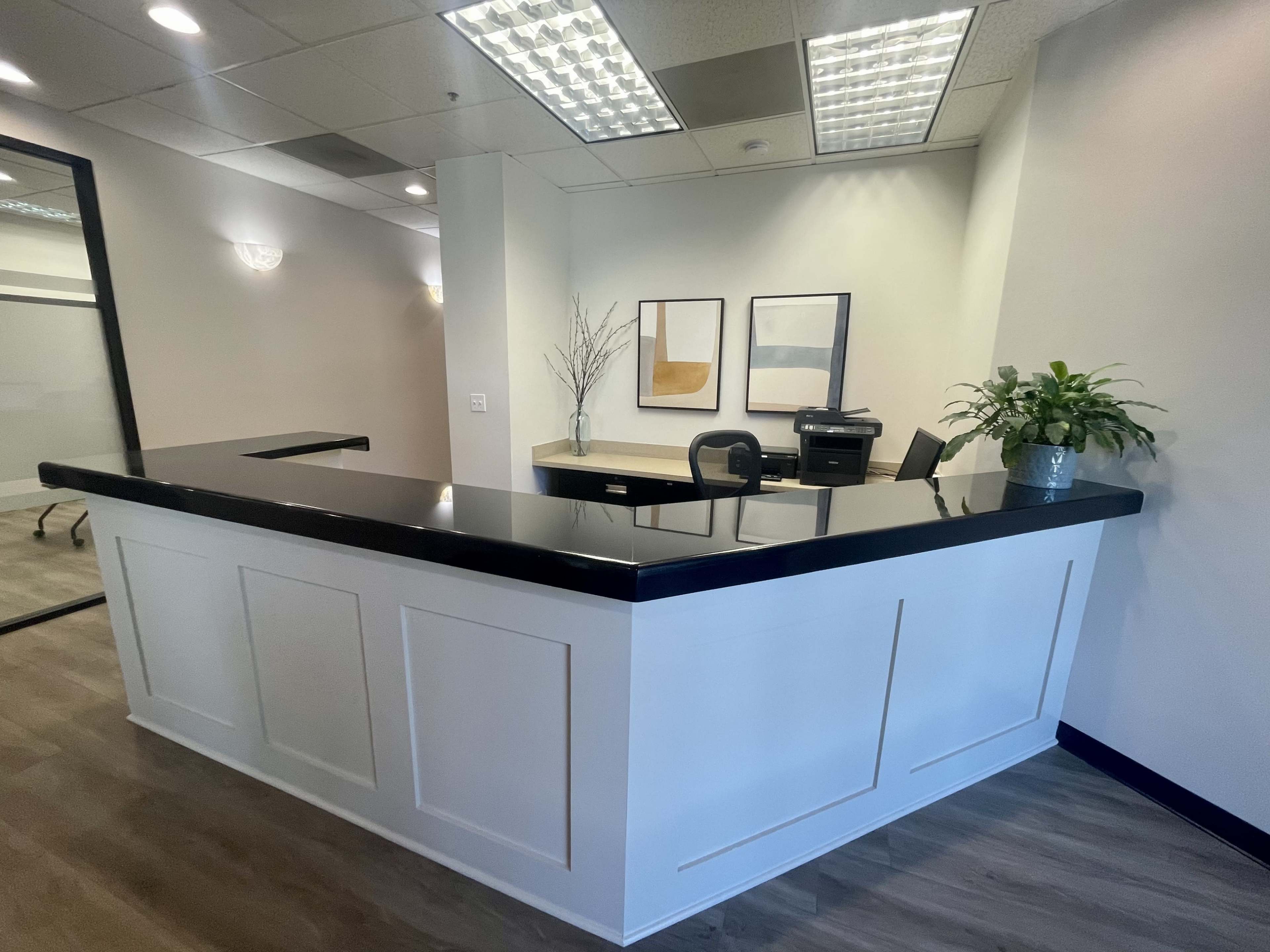 A modern reception area with a black countertop, white paneling, potted plants, and framed artwork on the walls.