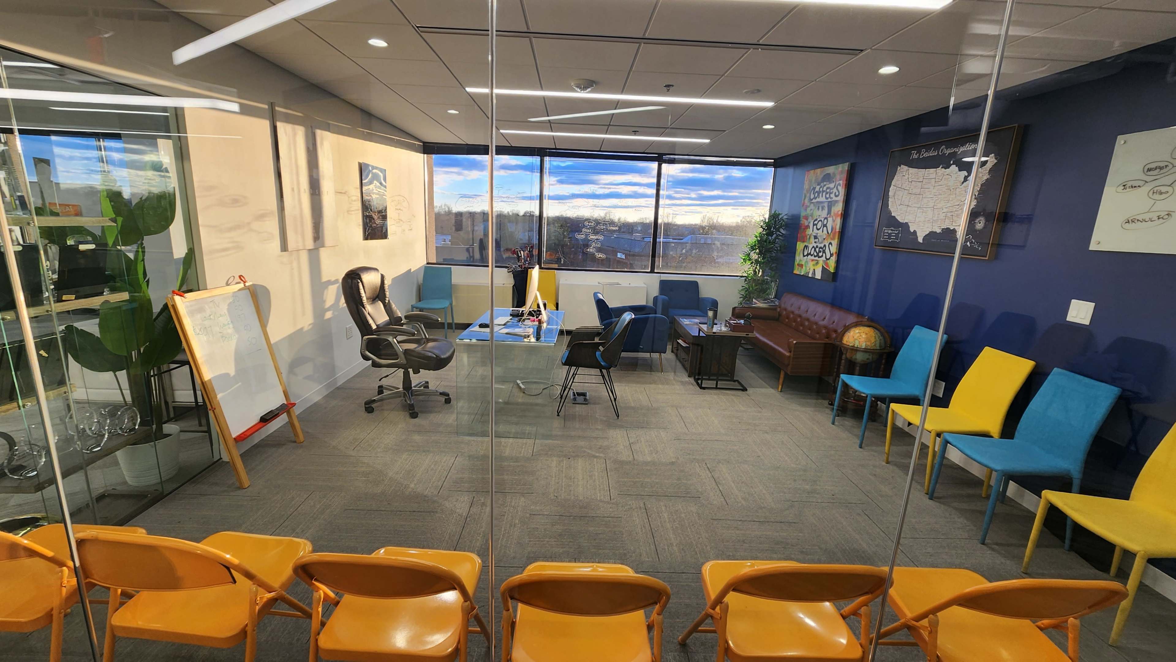 A modern conference room with glass walls, featuring a rectangular table, several chairs in bright colors, a large window with a view, and various decorations on the walls.
