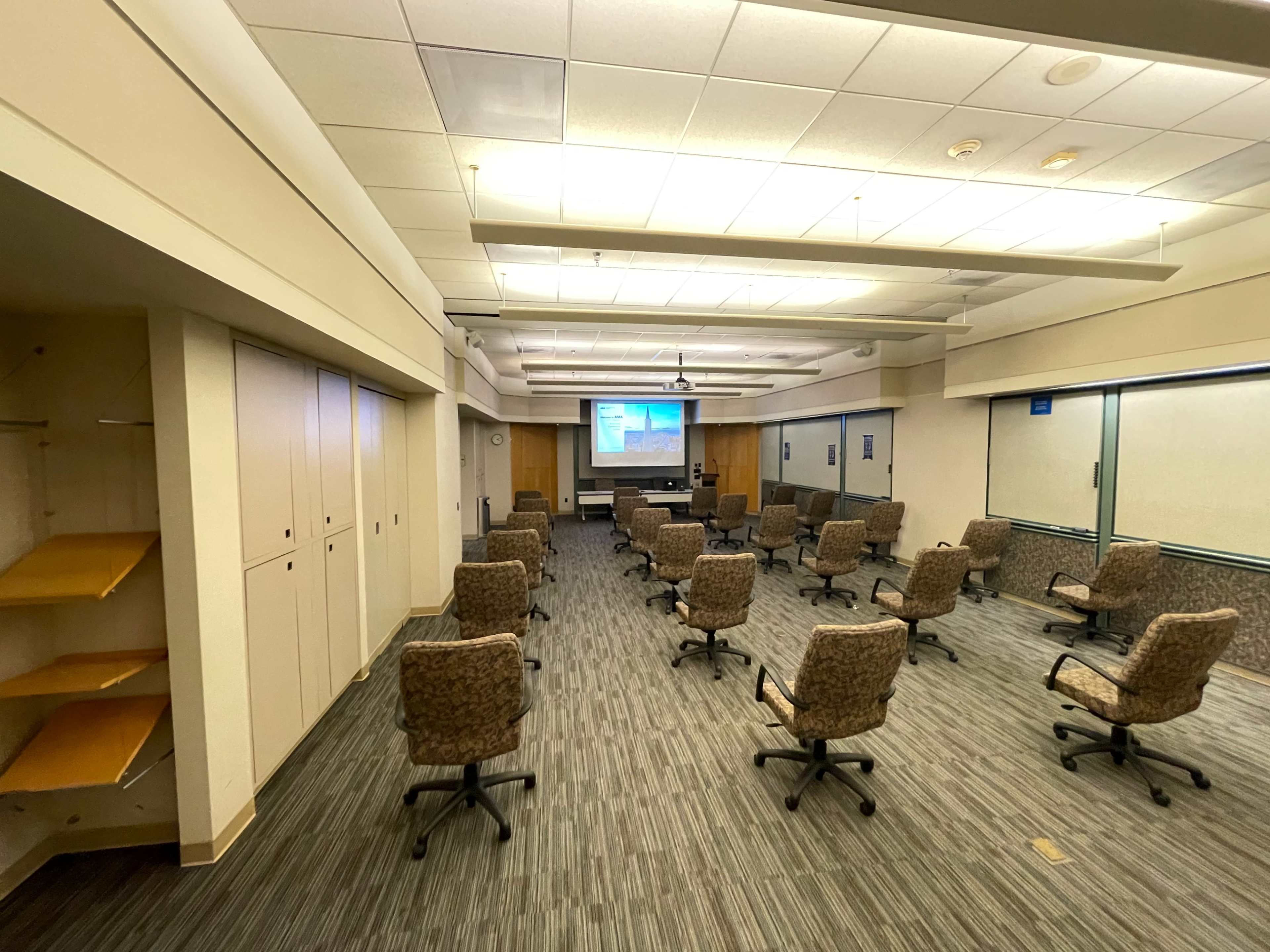 A conference room with rows of chairs positioned around a central presentation area and a projector screen at the front.