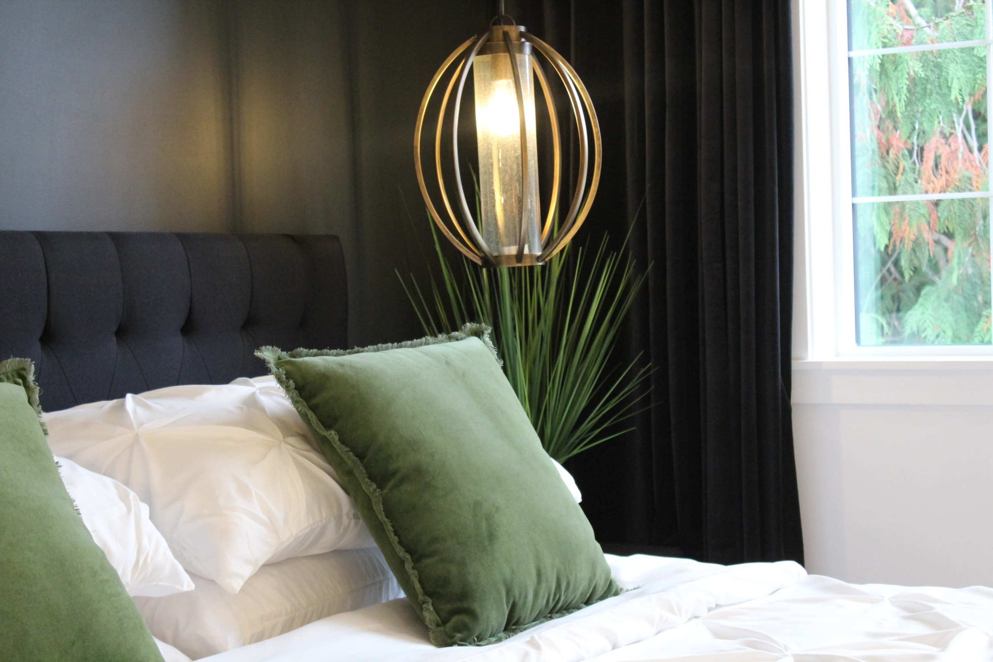 A neatly made bed with green pillows, a modern lamp, and a large window revealing greenery outside.