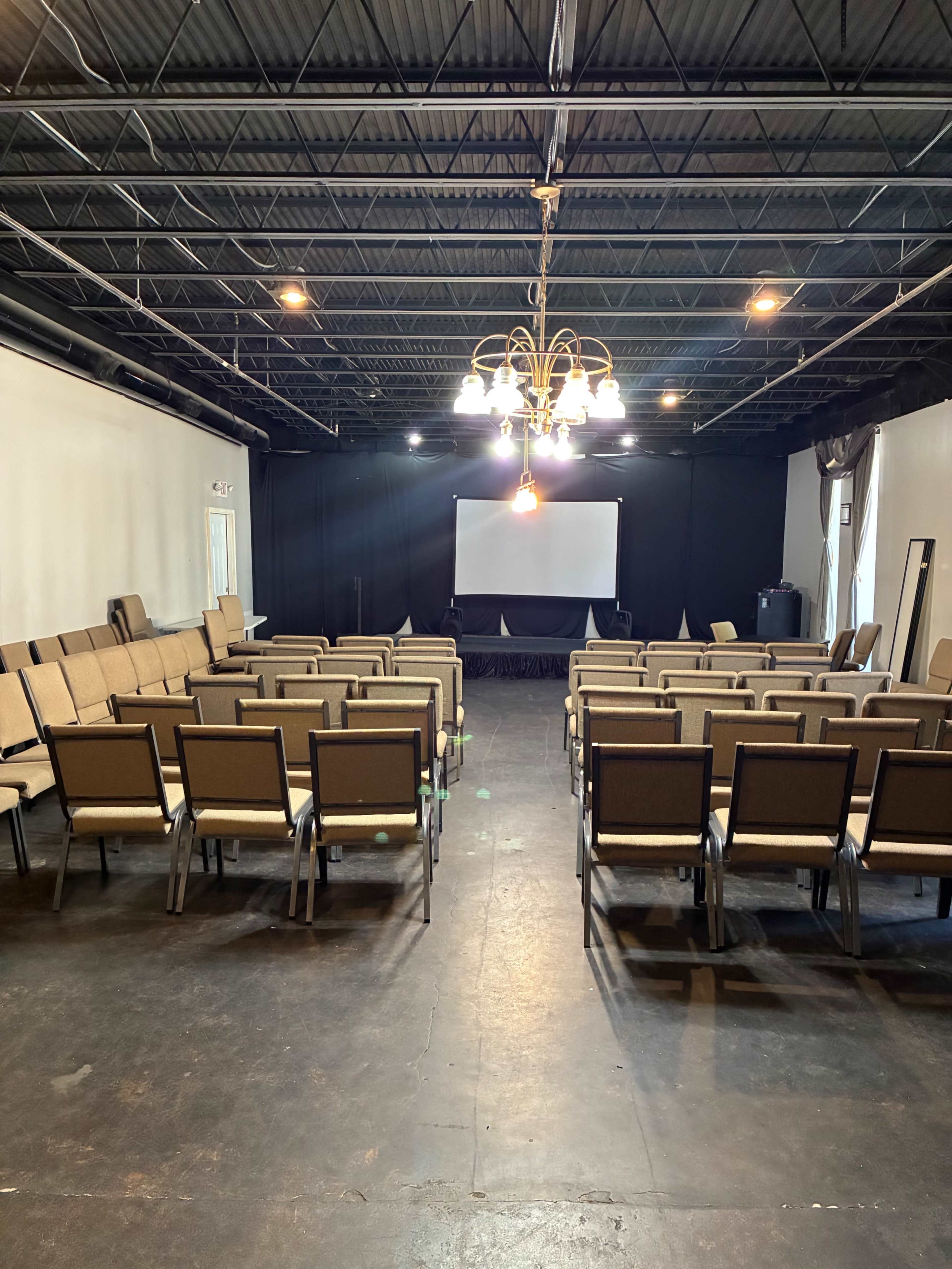 The image shows a sparsely furnished indoor auditorium with rows of chairs facing a projection screen at the front.
