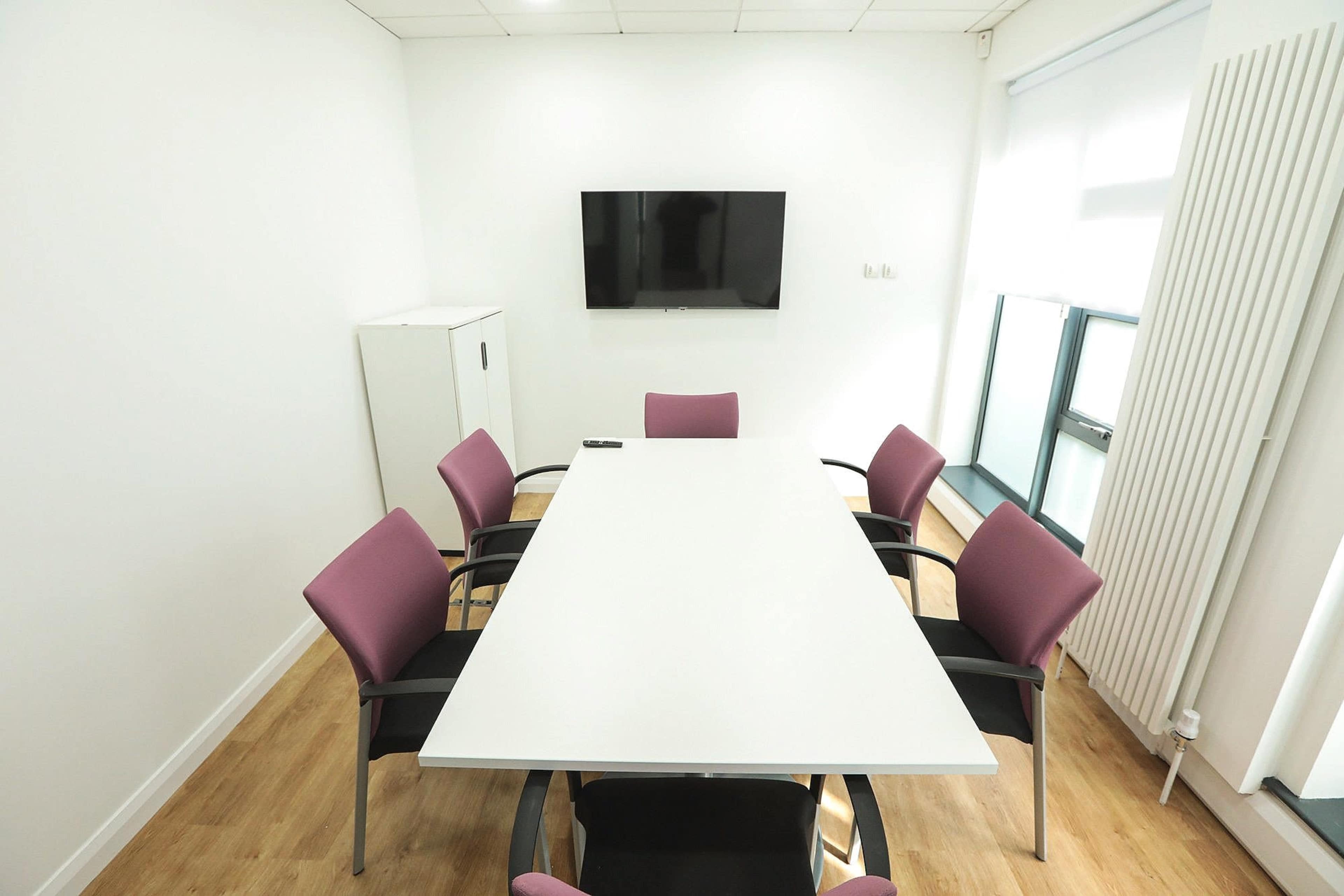 A conference room features a large white table surrounded by eight purple chairs and a wall-mounted television.