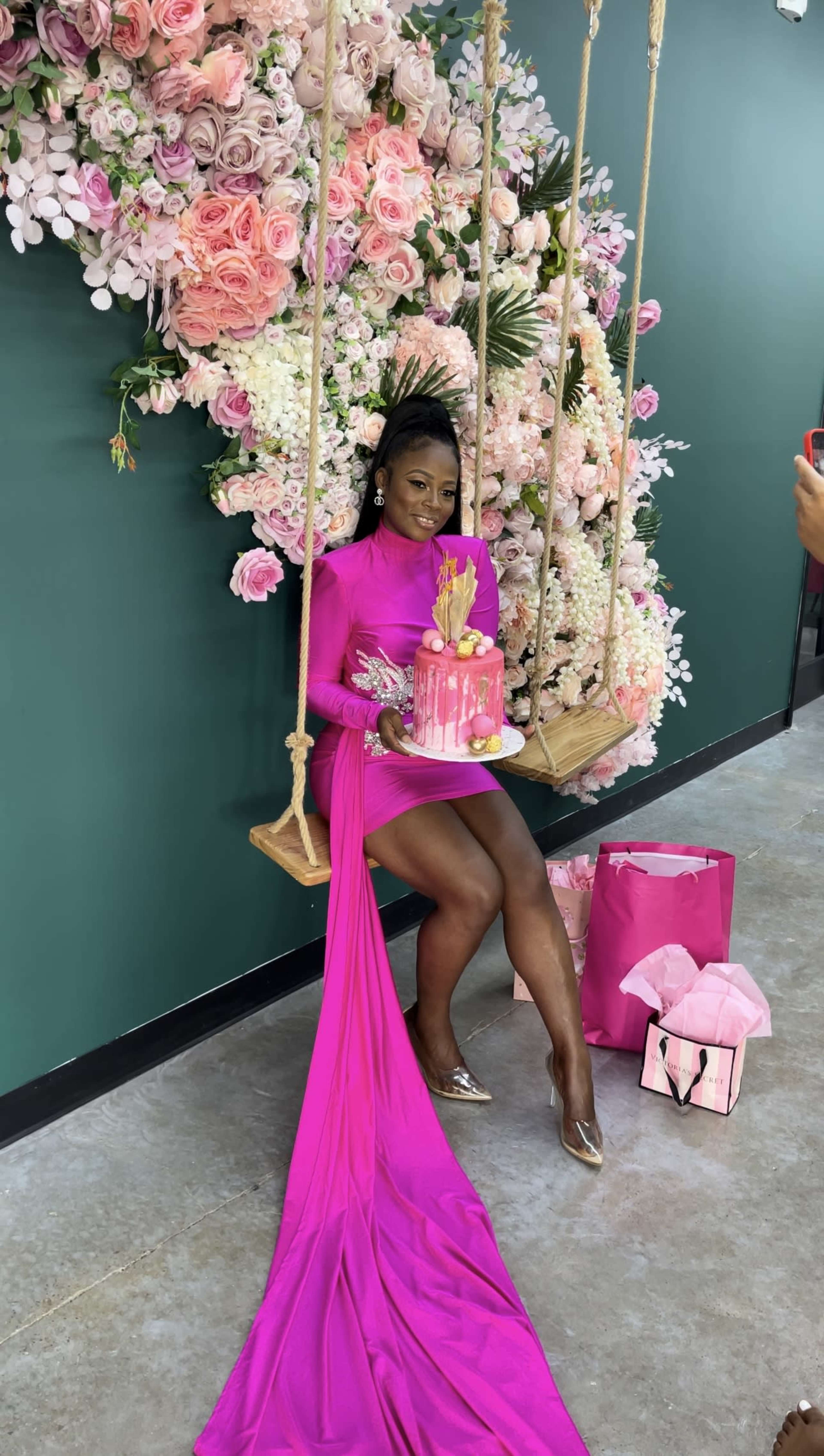 A woman in a pink dress poses on a swing adorned with a floral backdrop while holding a cake.