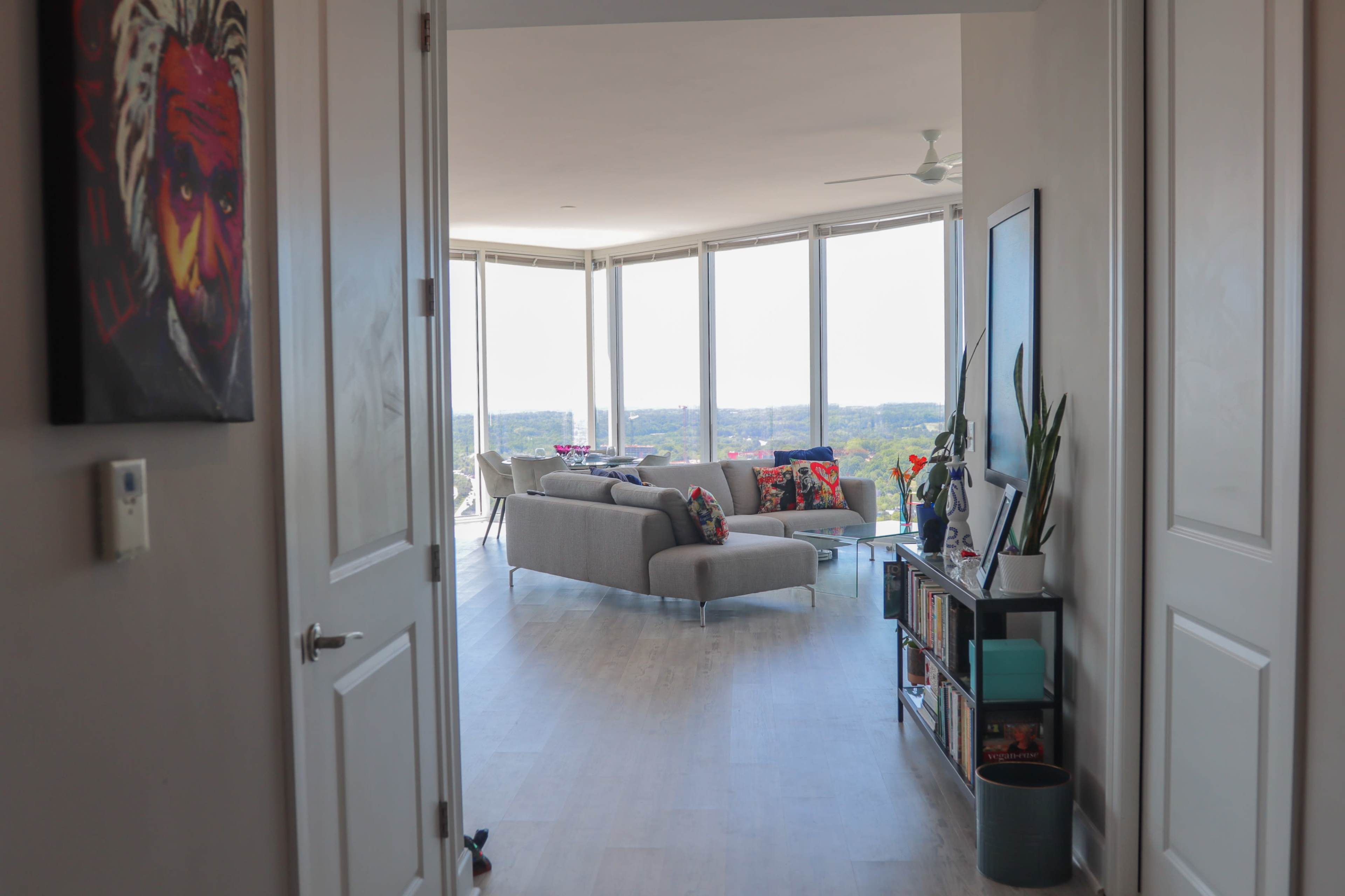 A hallway leads to a bright living room filled with large windows overlooking a scenic view.