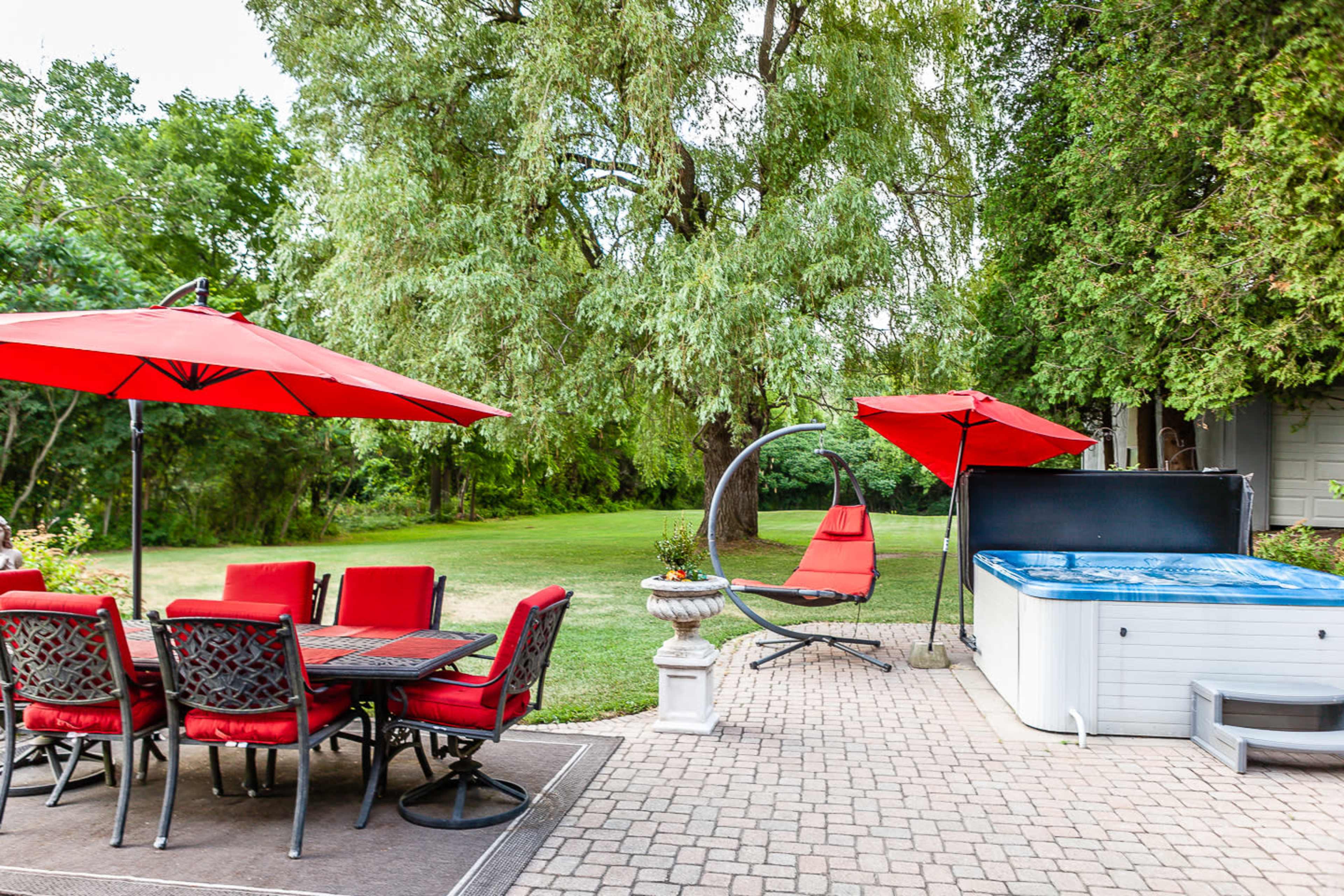 A backyard scene with a dining table set, red umbrellas, a hanging chair, and a hot tub surrounded by greenery.