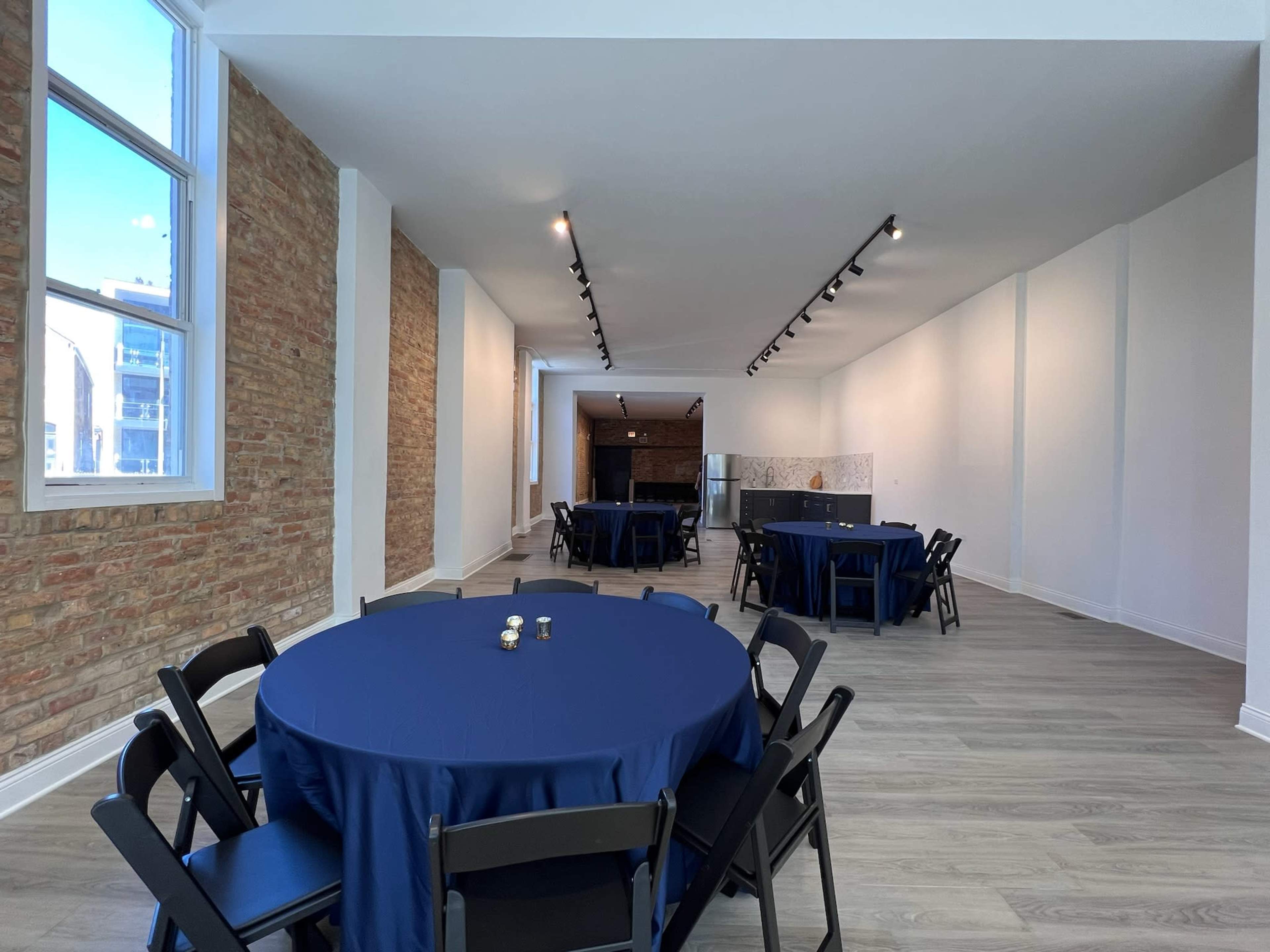 An empty event space featuring several round tables covered with navy tablecloths, surrounded by black chairs, and illuminated by ceiling lights.