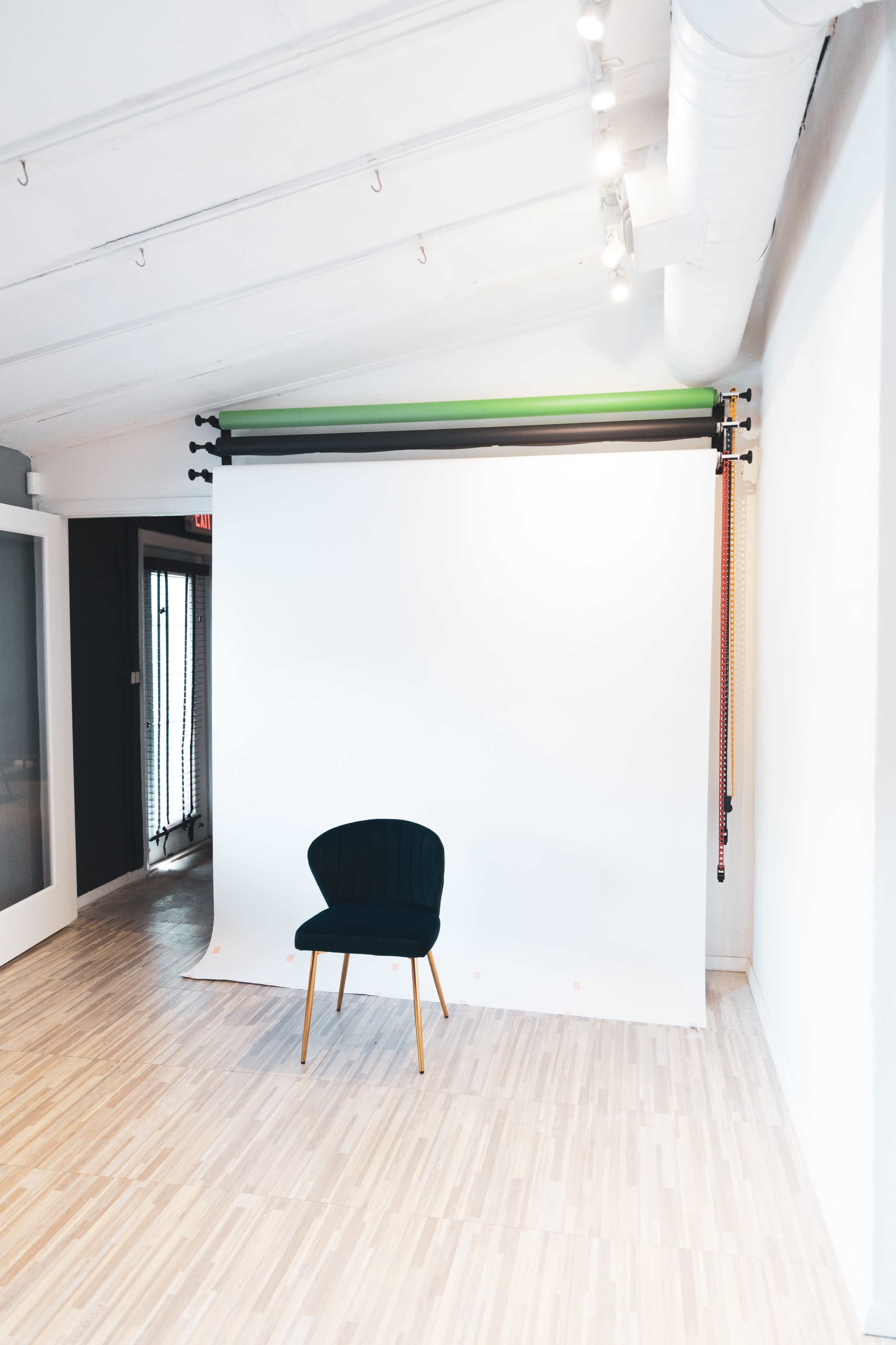 A green and black backdrop is set up in a bright room with a single chair positioned in front of a white screen.