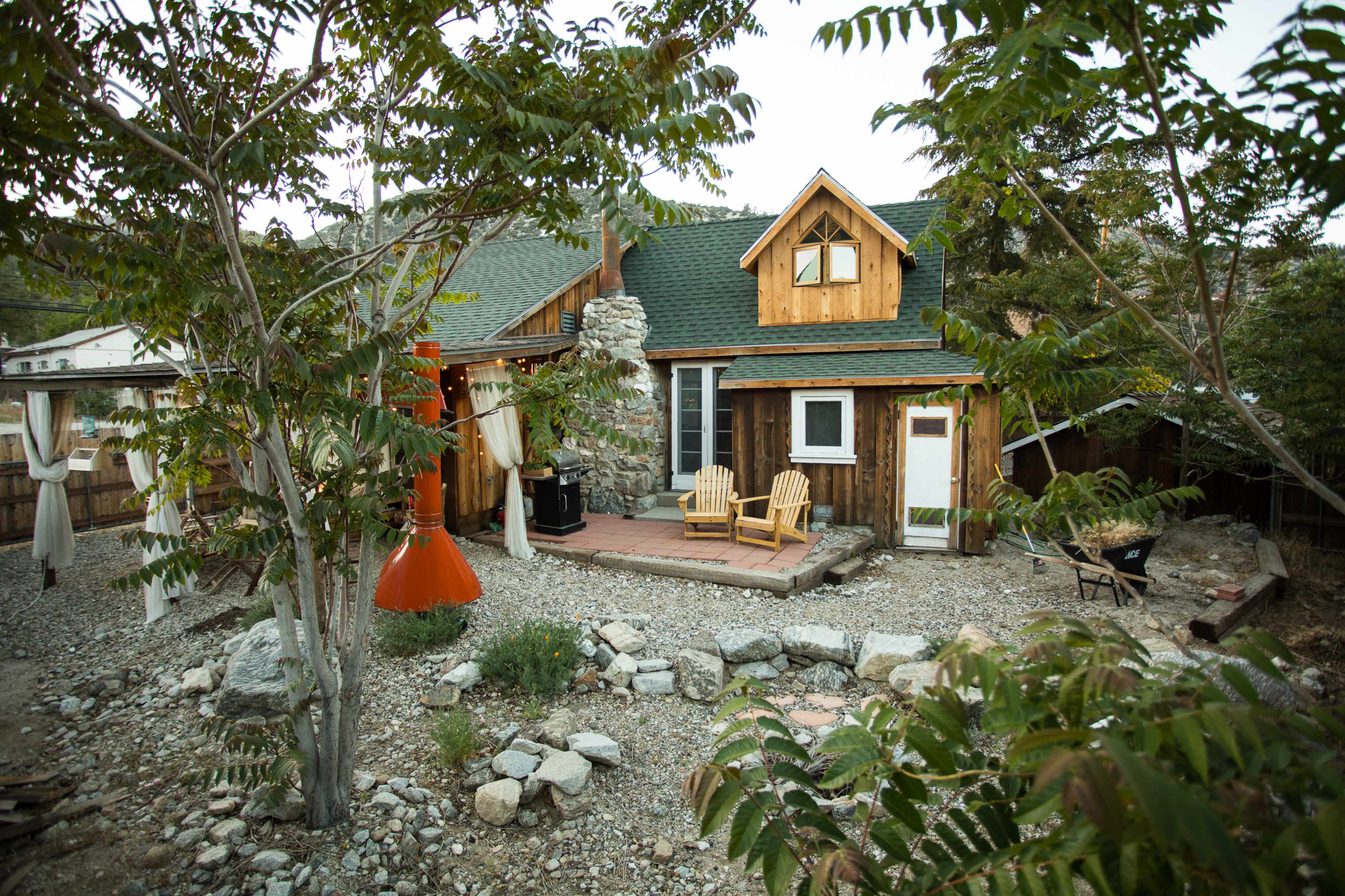 A wooden cottage with a stone chimney and a deck, surrounded by gravel and trees, sits in a rural setting.