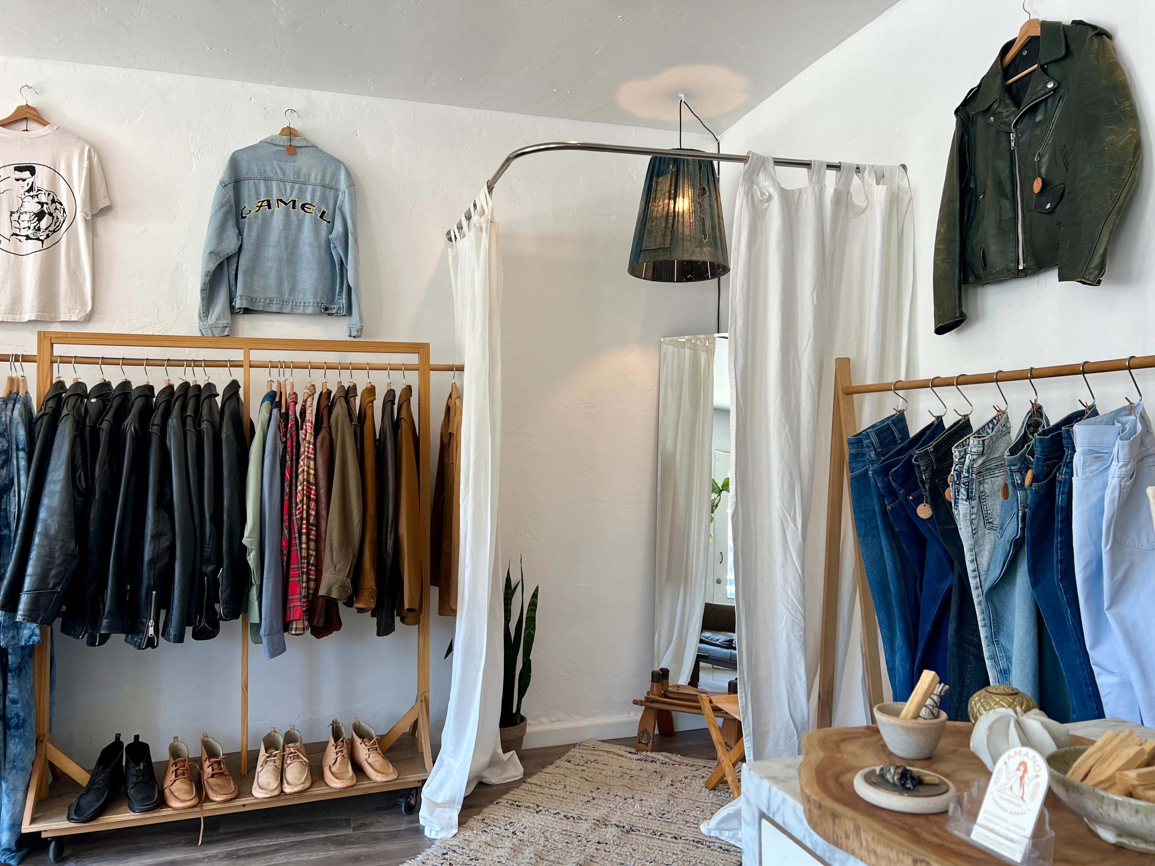 A clothing boutique featuring racks of jackets and jeans, hanging garments on the walls, and a small seating area with a plant and decorative items.