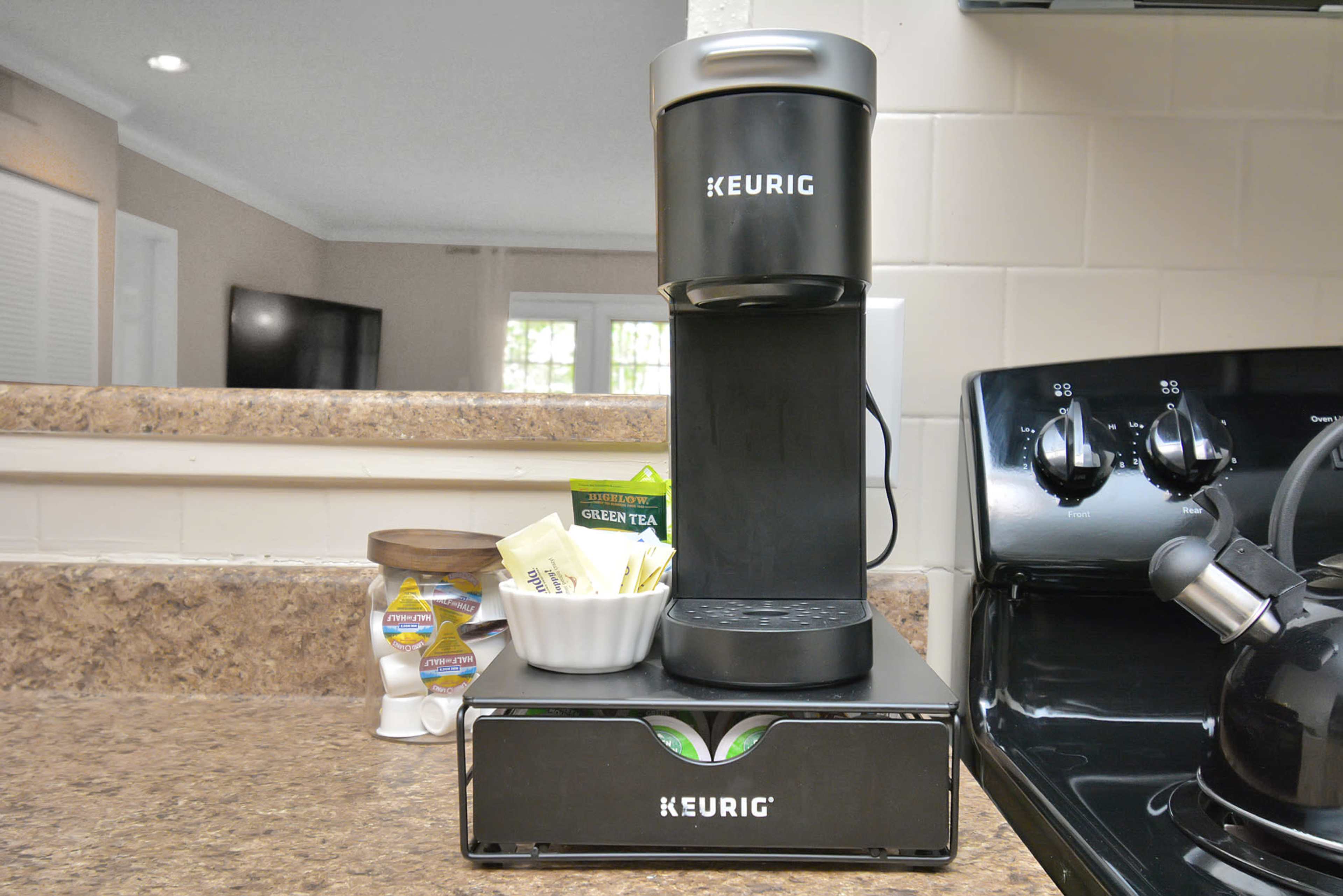 A Keurig coffee maker is placed on a countertop next to a drawer holding coffee pods and a bowl with tea bags.