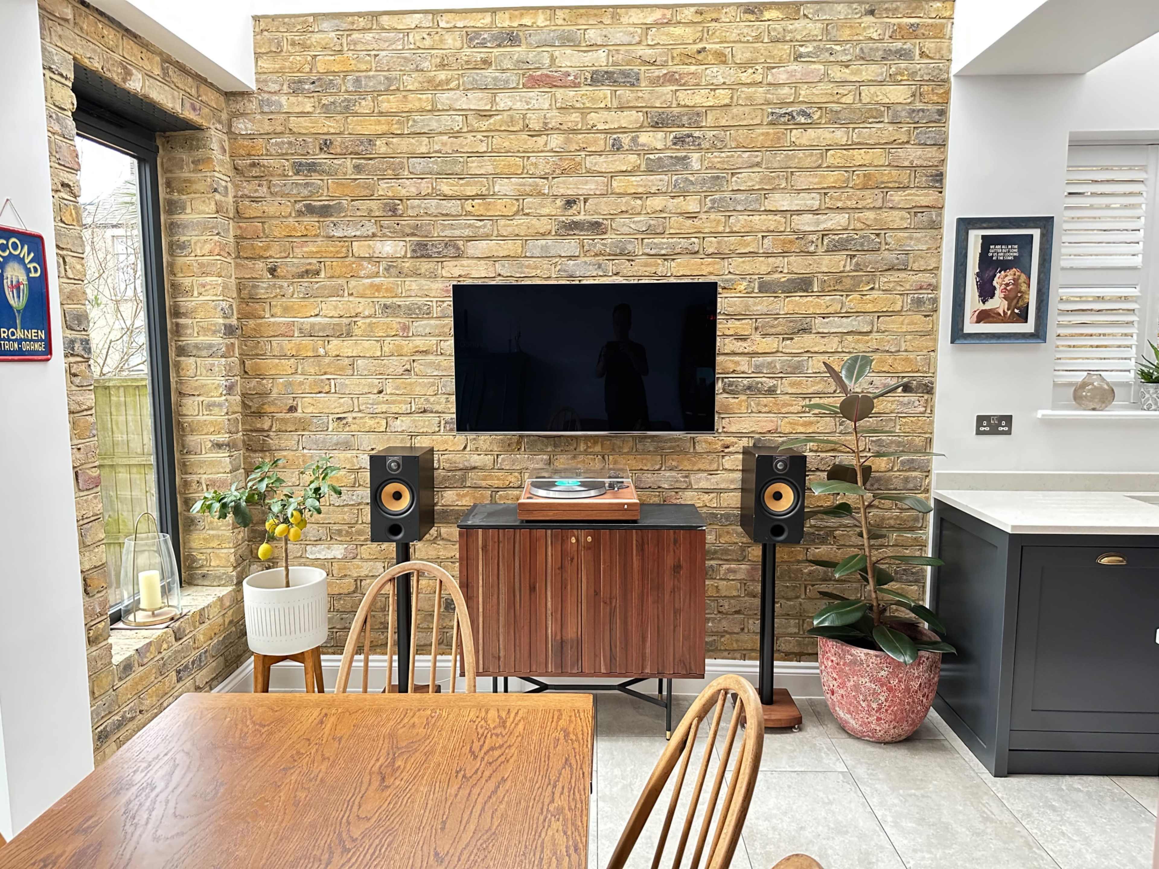 The image shows a modern living space featuring a brick wall, a television mounted above a wooden cabinet with a turntable, and speakers positioned on either side of the cabinet.