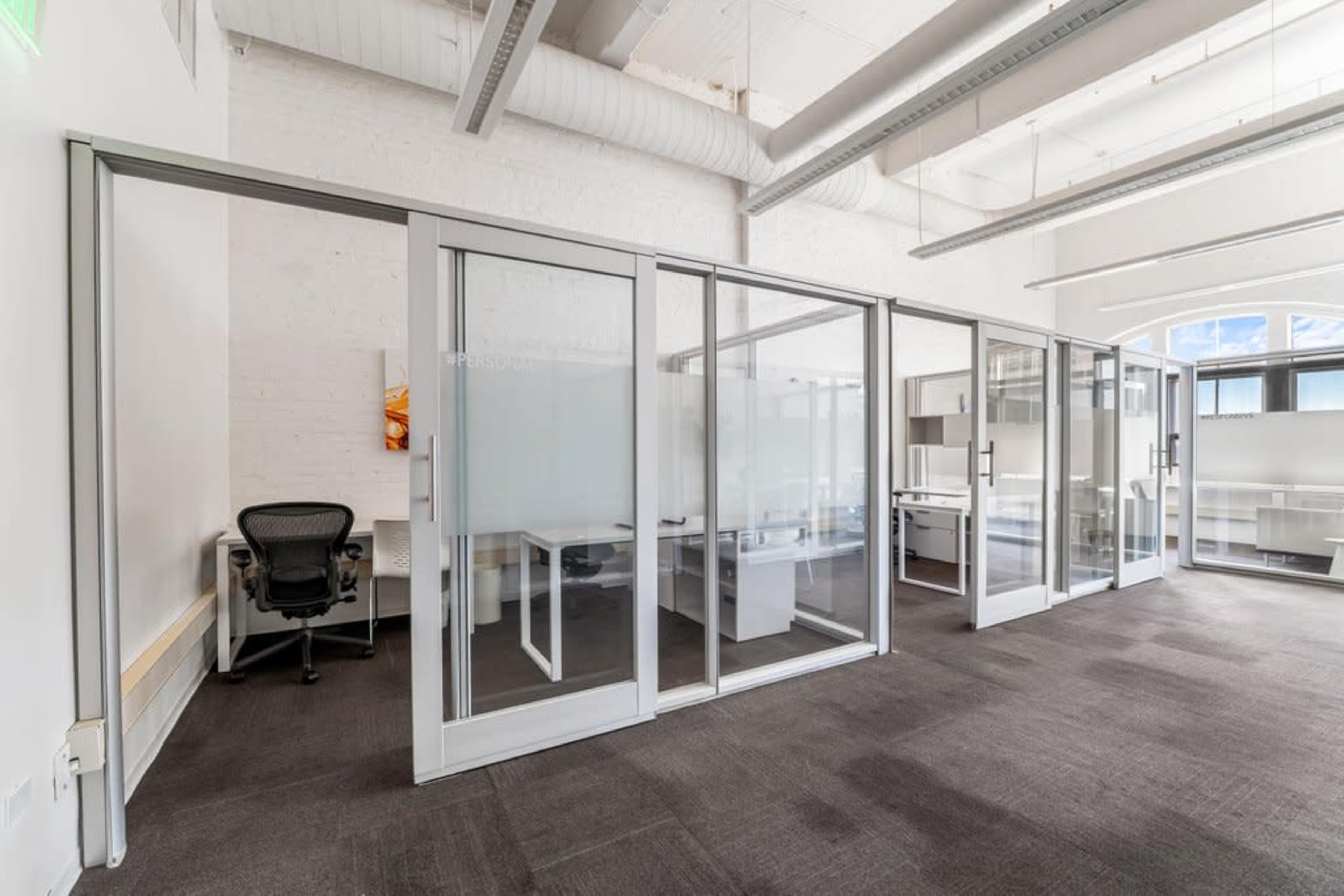The image shows a modern office space with glass-walled meeting rooms and a single black office chair.