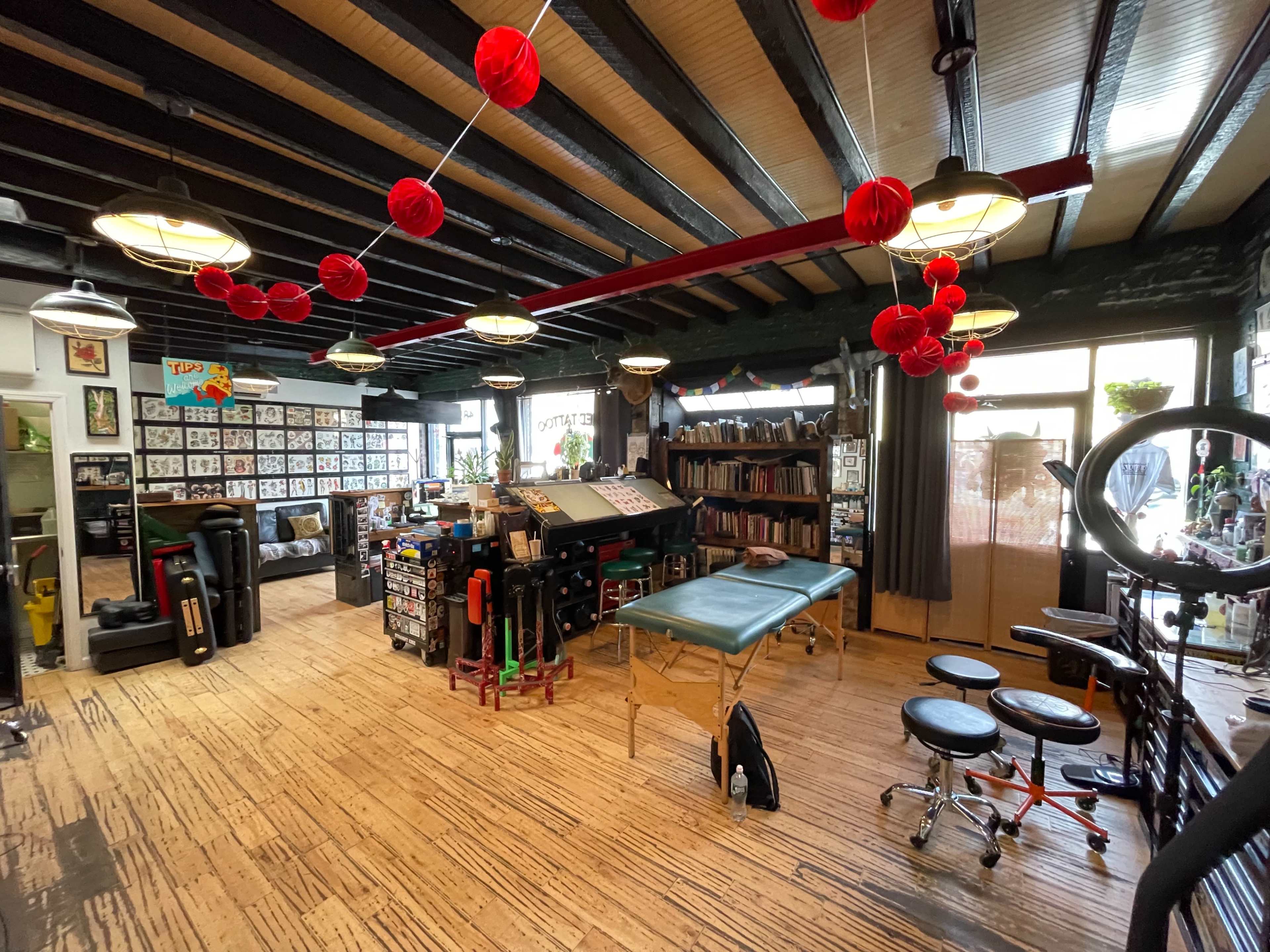 The image shows a tattoo shop with wooden beams, tattoo stations, art displays on the walls, and decorative red lanterns hanging from the ceiling.