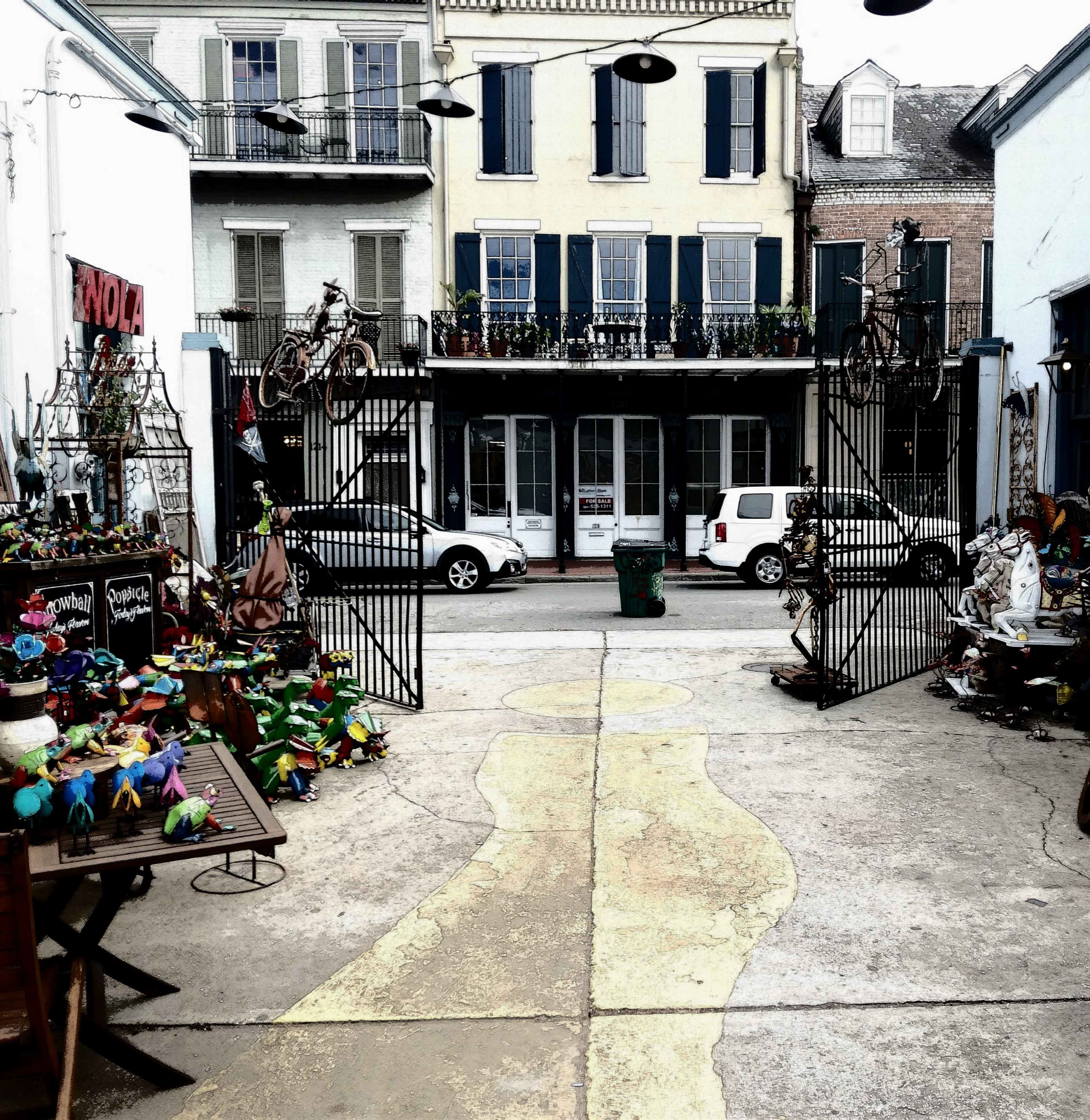 A courtyard features a pathway lined with colorful decorations and leads to a set of black gates in front of a yellow building with multiple balconies.