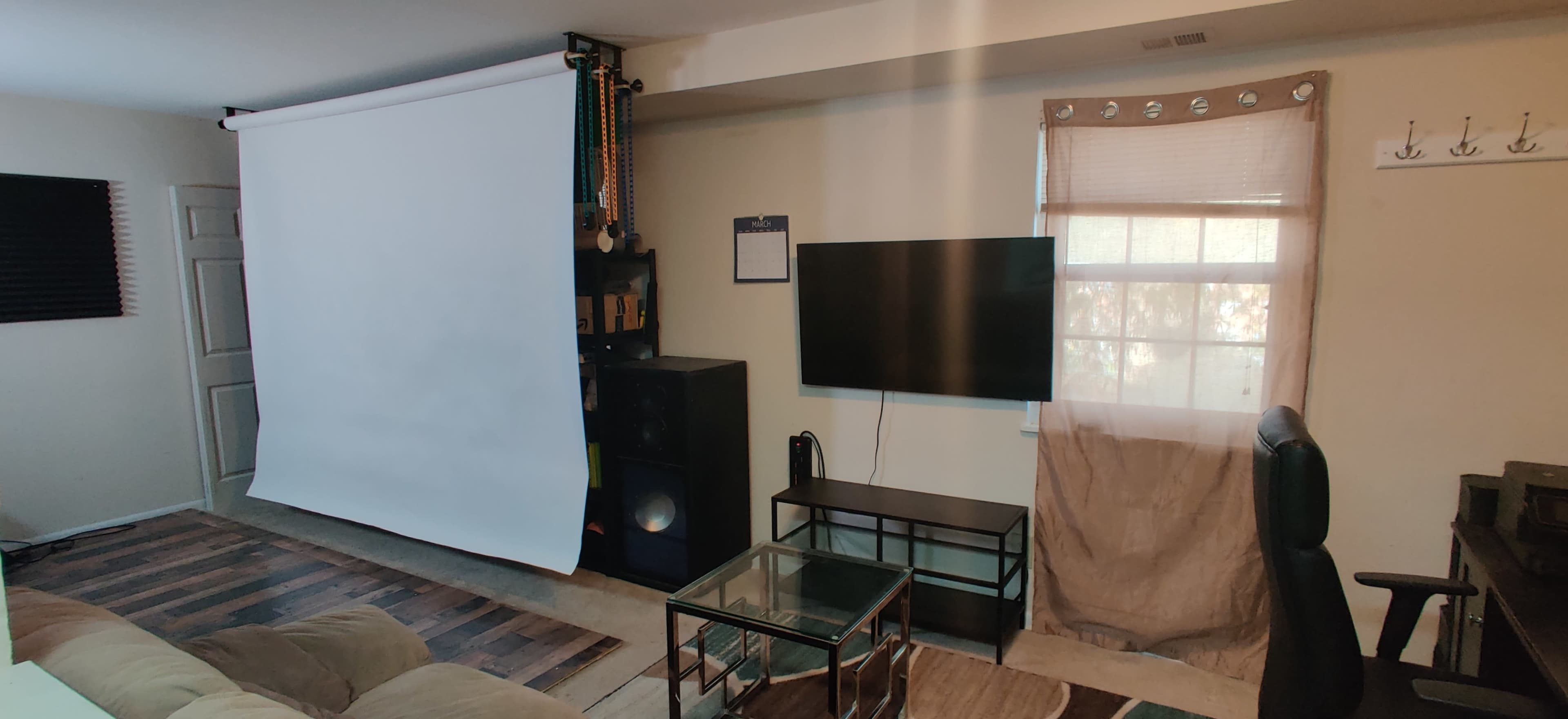 A living room features a projection screen, a television mounted on the wall, a glass coffee table, and a sofa, with a window partially covered by a curtain.