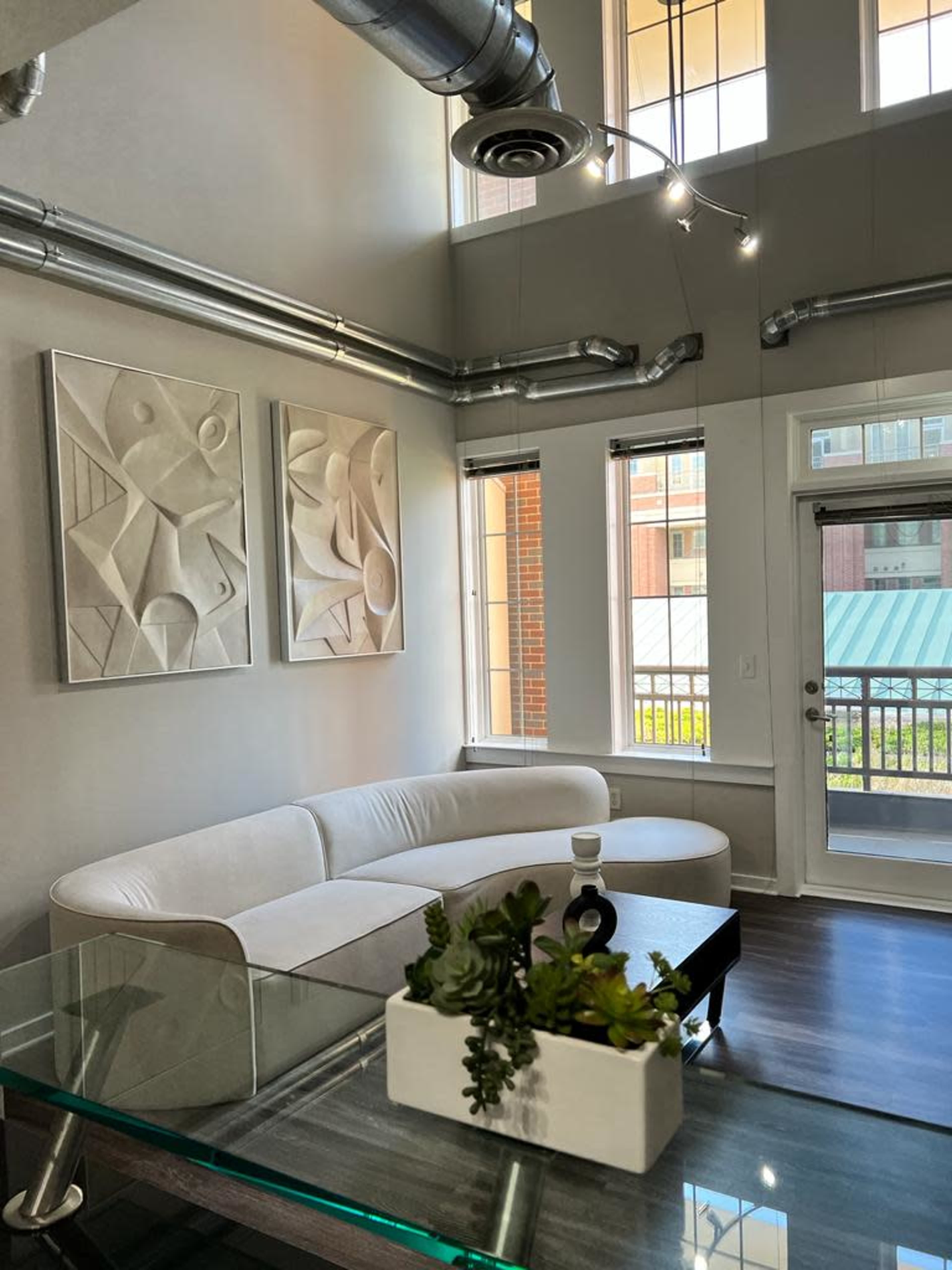 A modern living room with a curved white sofa, abstract wall art, large windows, and a glass table with a small plant arrangement.