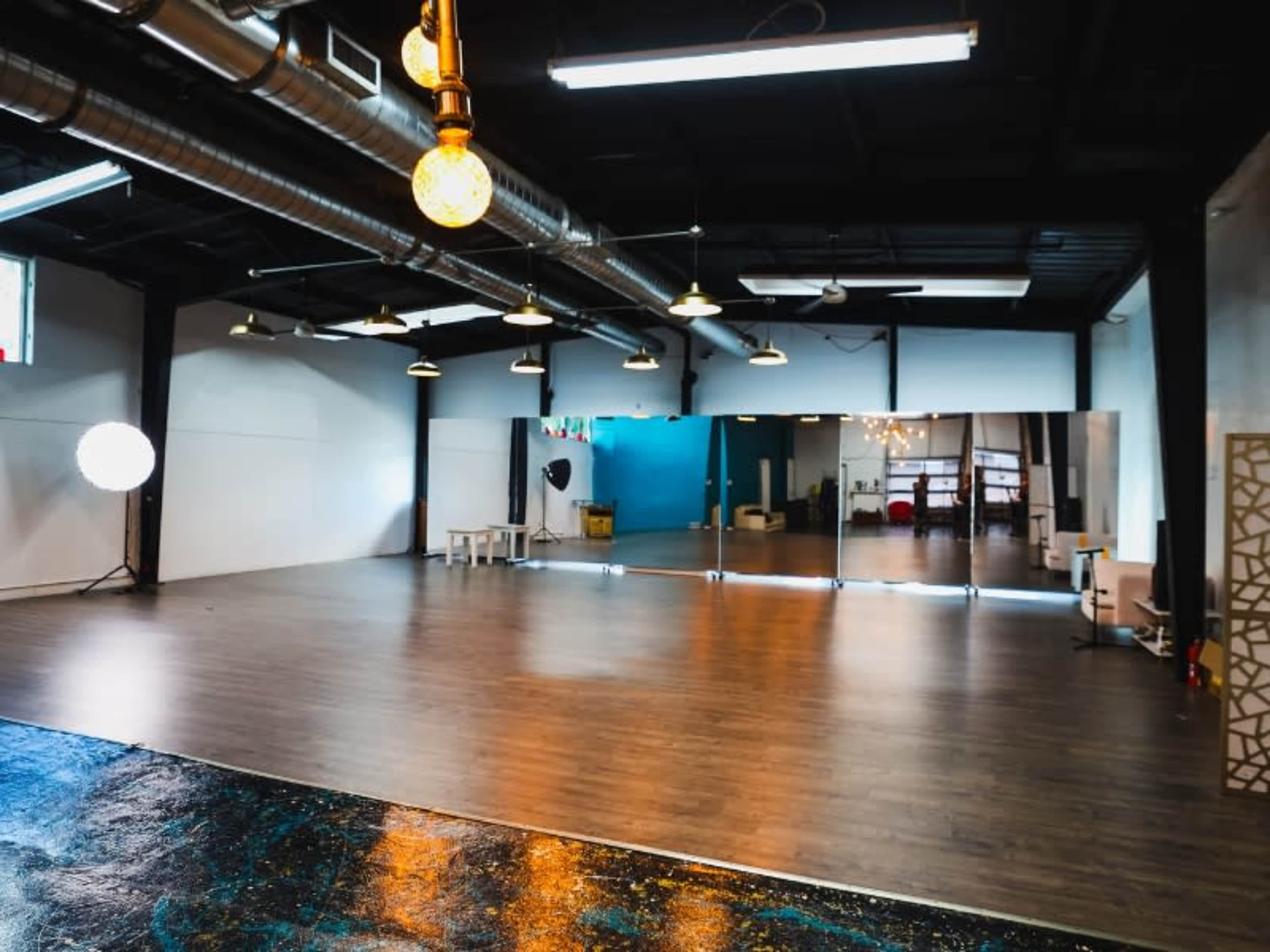 A spacious dance studio with wooden flooring, large mirrors along one wall, and industrial lighting.
