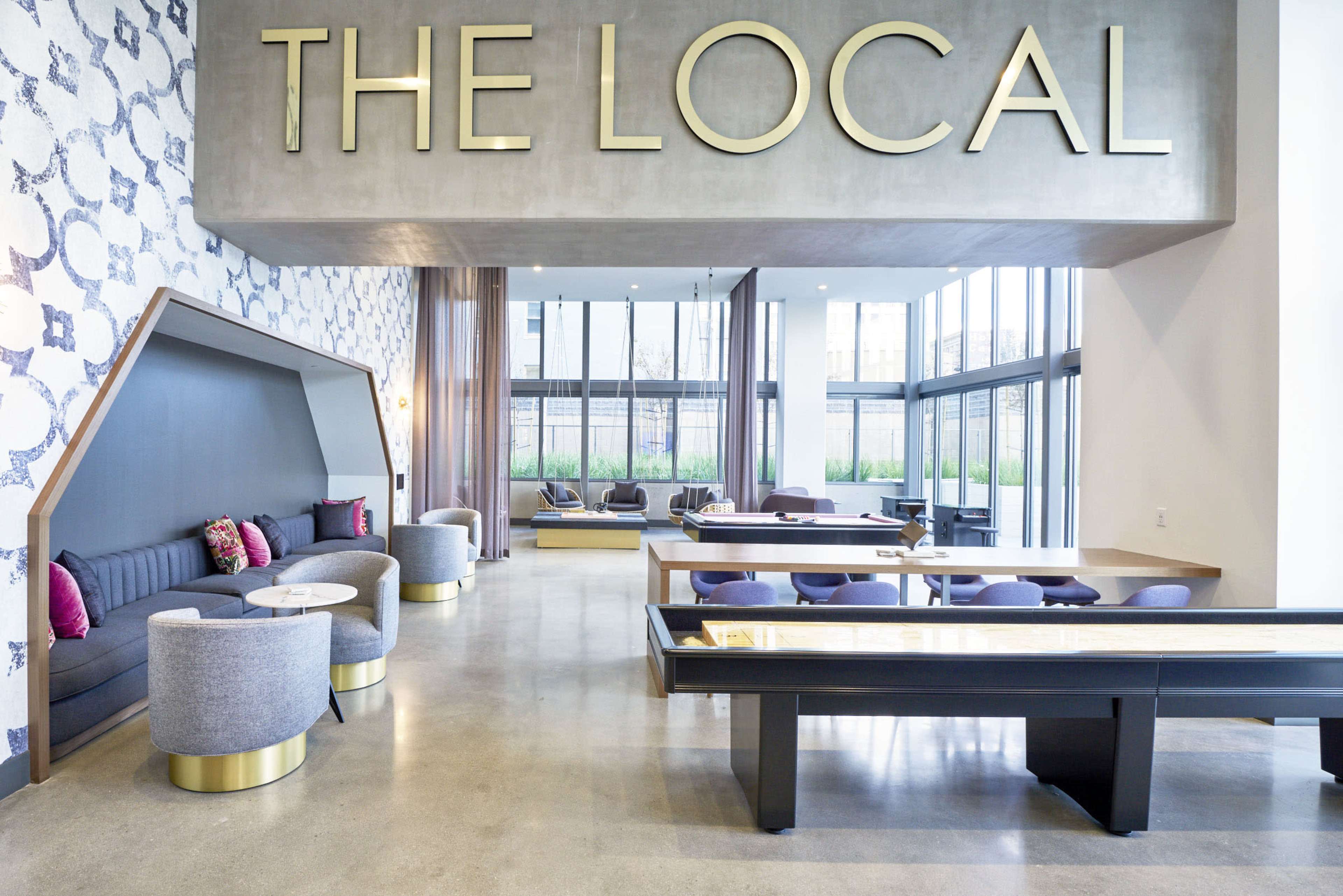 A modern lobby area with a large sign reading "THE LOCAL," featuring seating arrangements and large windows that allow natural light to enter.