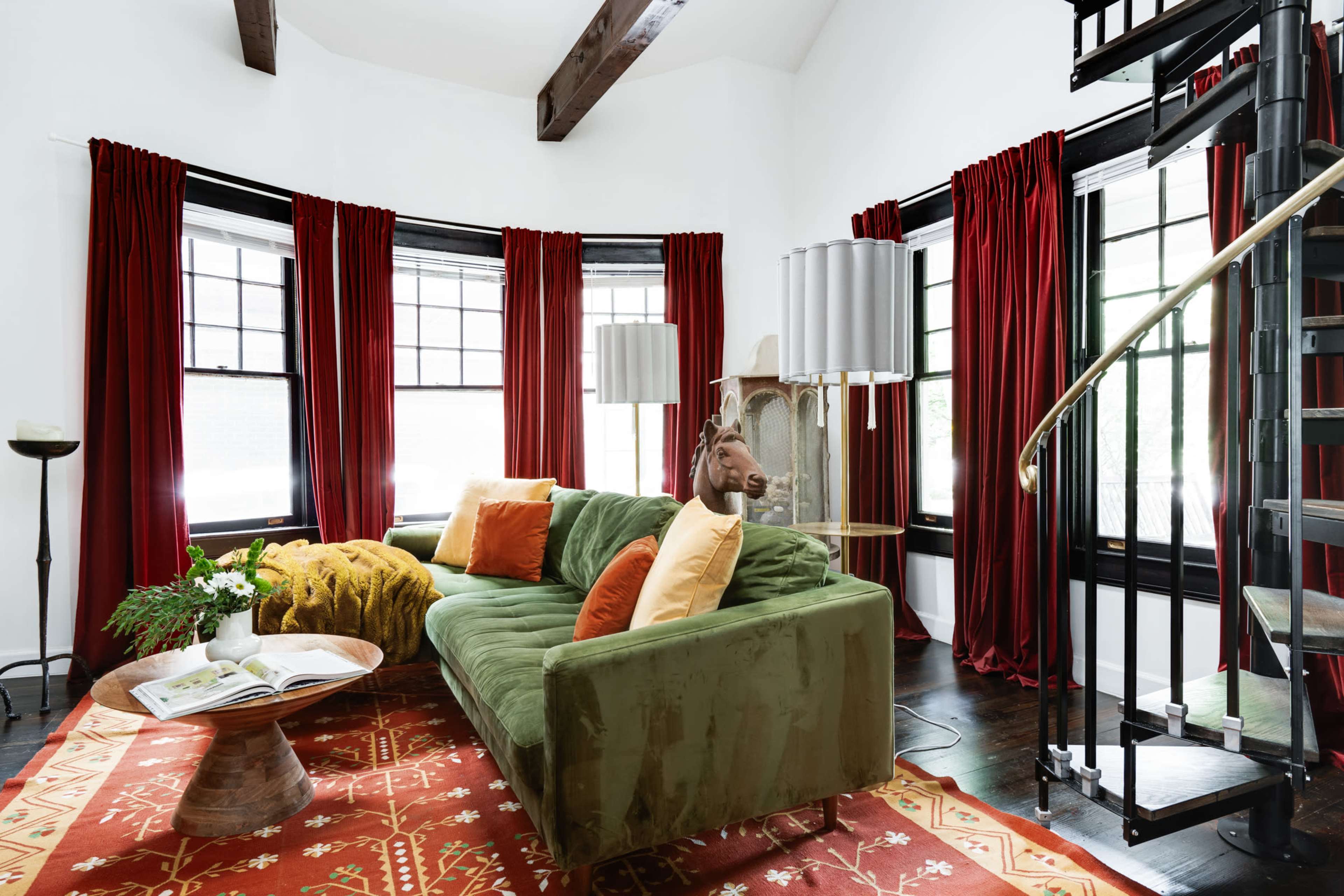The living room features a green sofa with colorful cushions, a round wooden coffee table, and large windows dressed with red curtains, alongside a spiral staircase.