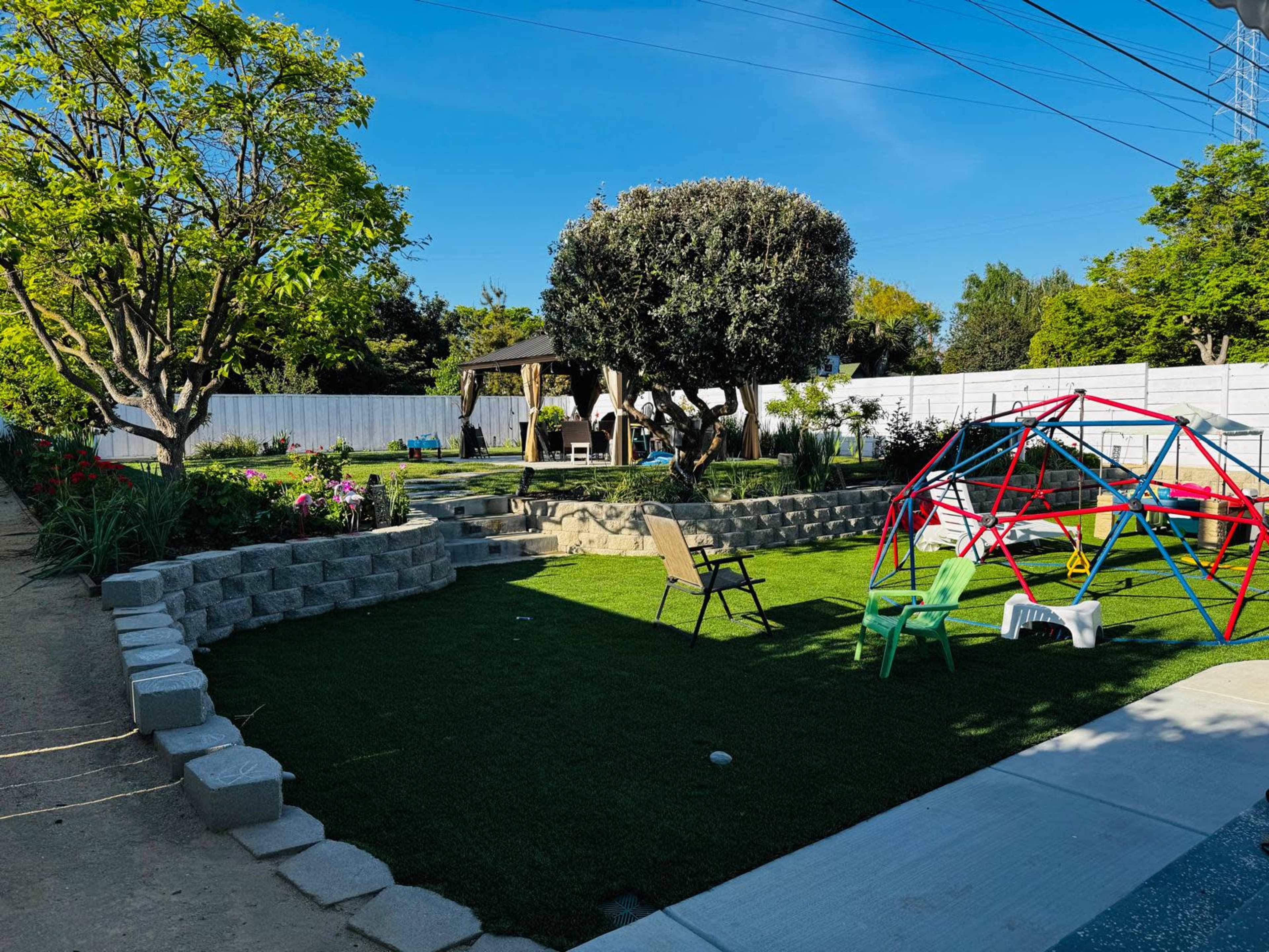 The image shows a landscaped backyard featuring a grassy area with a play structure, lounge chairs, trees, and a shaded seating area.