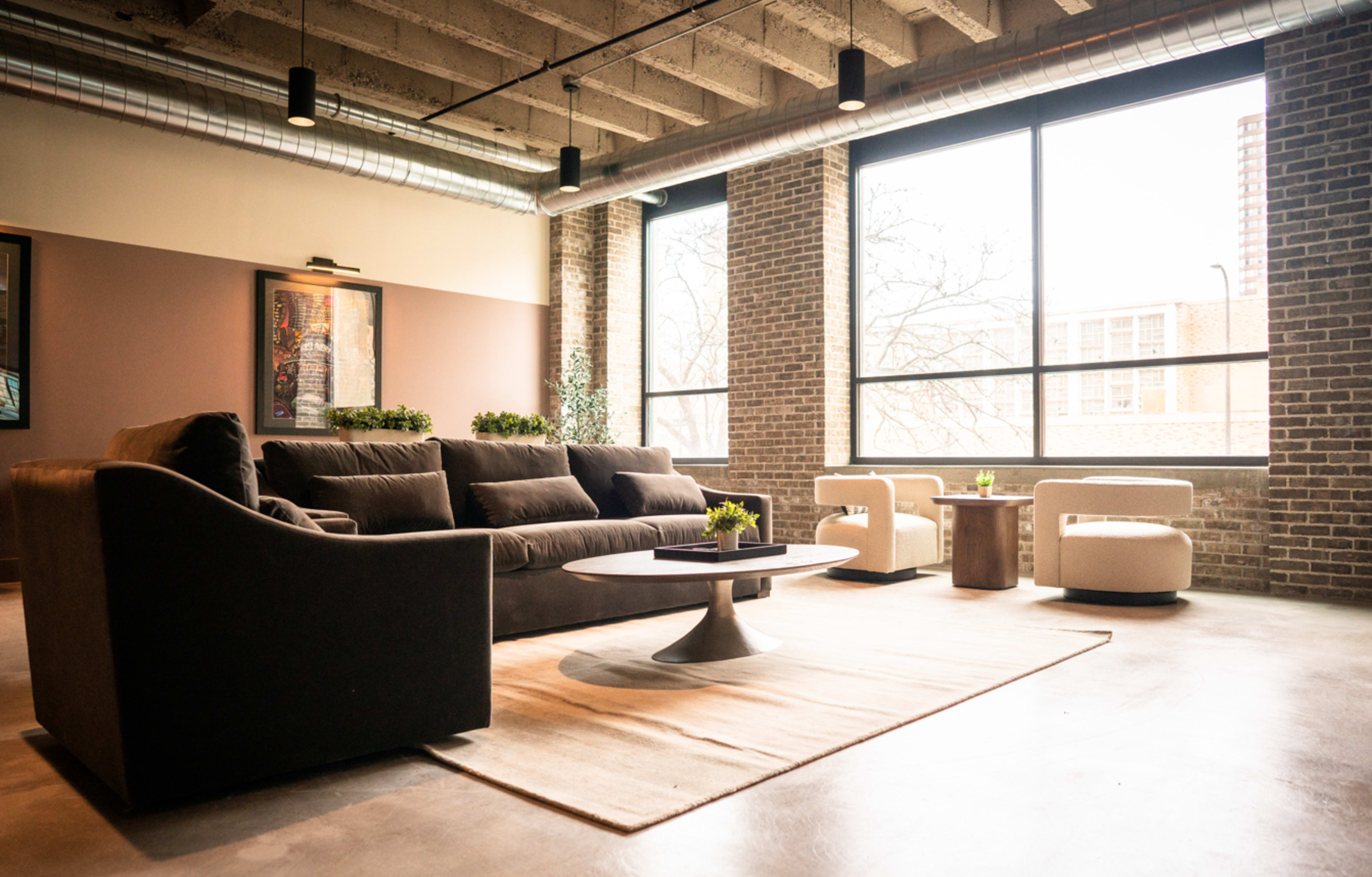 The image shows a modern lounge area with a dark sofa, light chairs, a round coffee table, and large windows allowing natural light to enter the space.