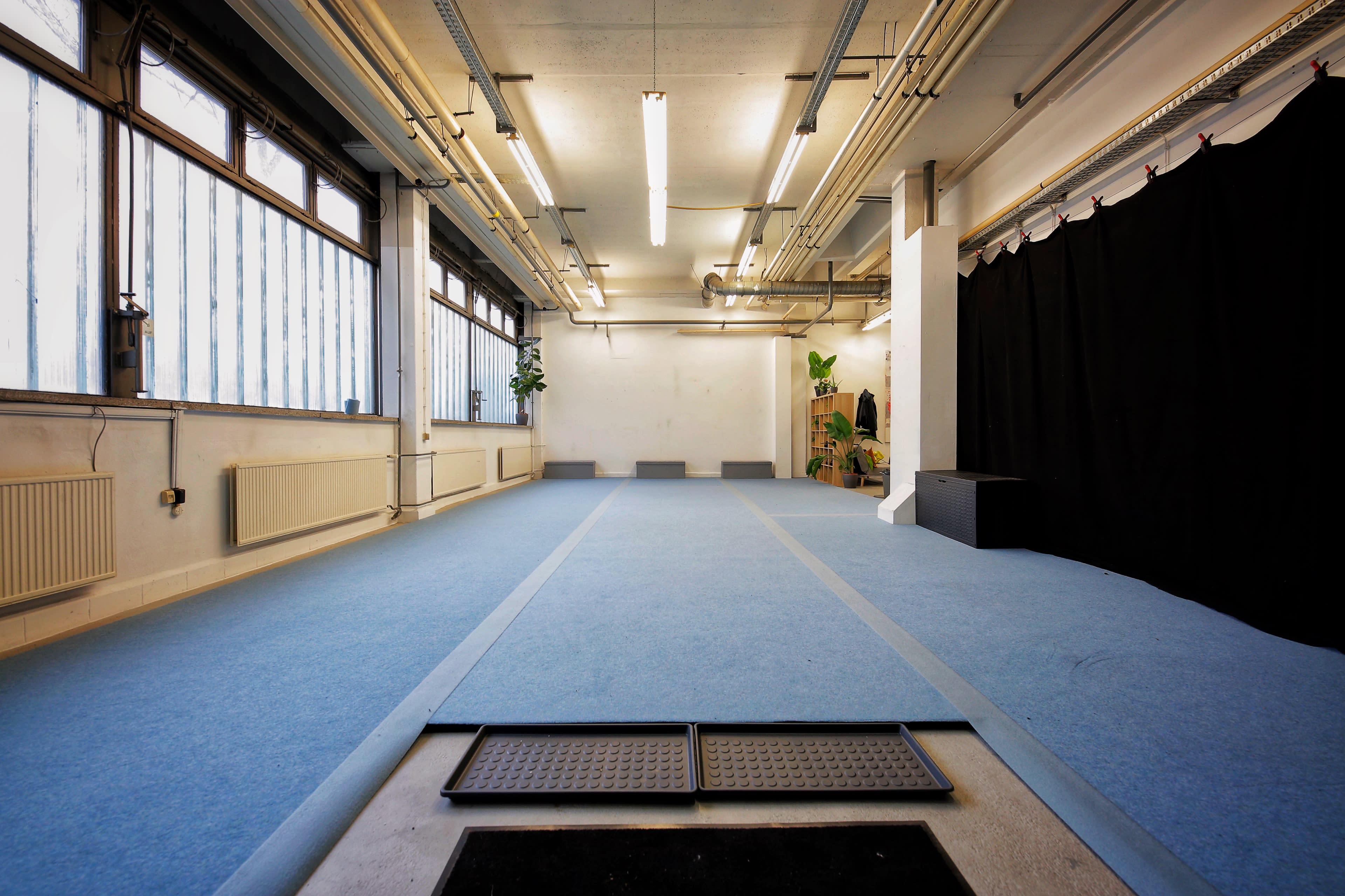 The image shows a spacious room with blue carpet flooring, large windows along one wall, and a black curtain at the far end.