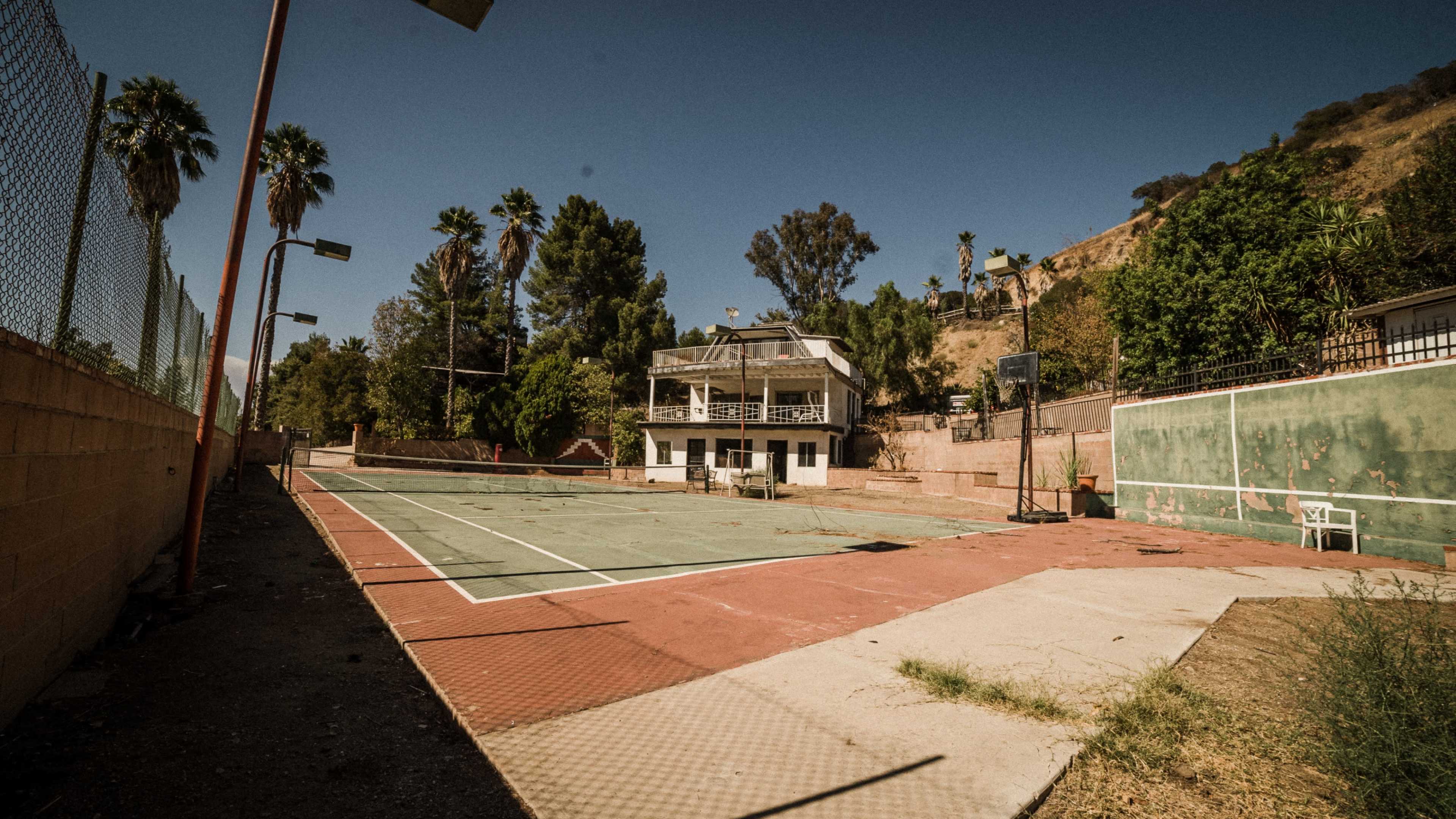 An abandoned tennis court with a dilapidated building in the background, surrounded by palm trees and overgrown vegetation.