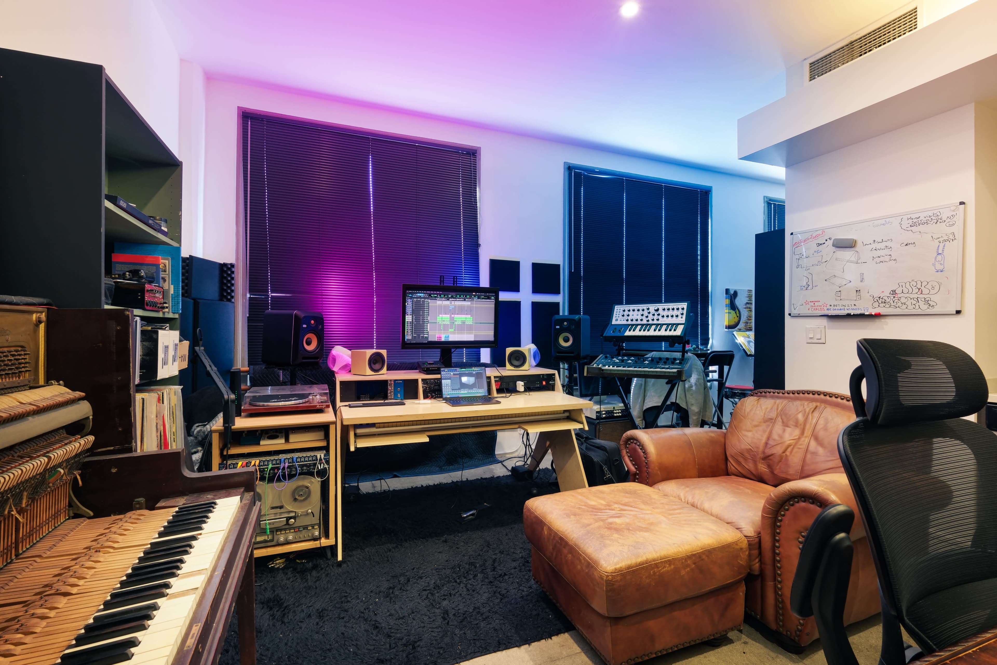 A music production studio contains a computer setup with monitors, various musical instruments, and a leather chair in a well-lit room with dark window blinds.