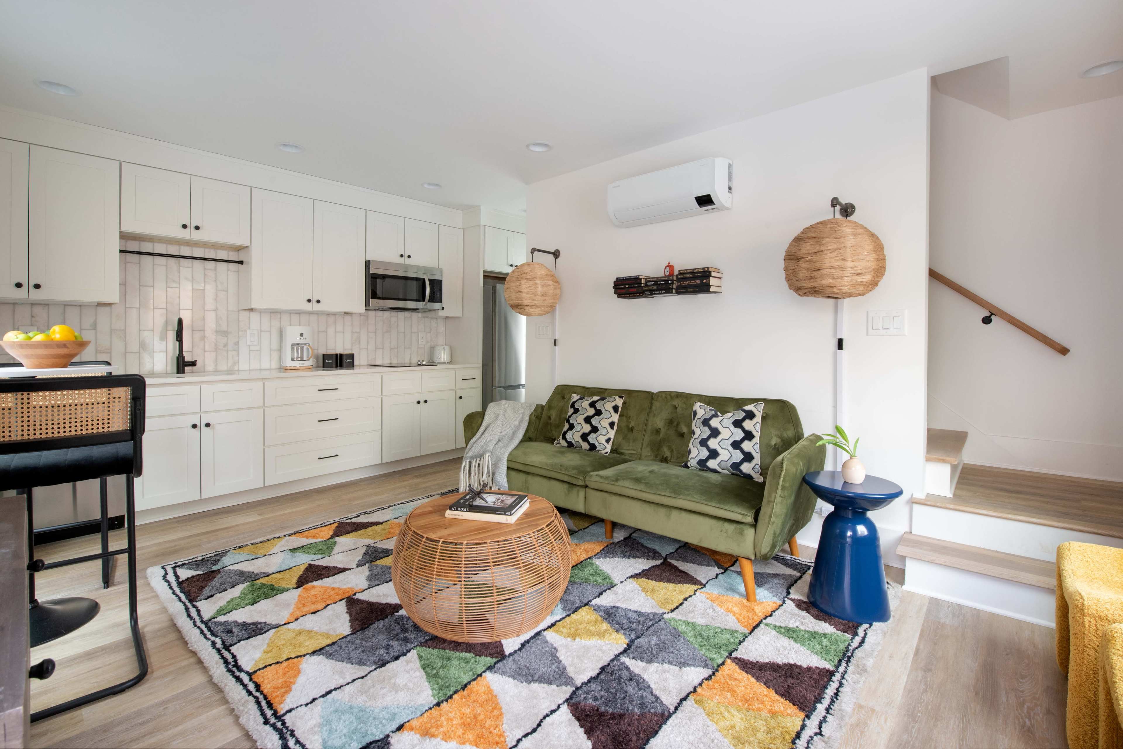 The image shows a modern living space featuring a green sofa, a patterned rug, a wooden coffee table, and a kitchen area with white cabinets.