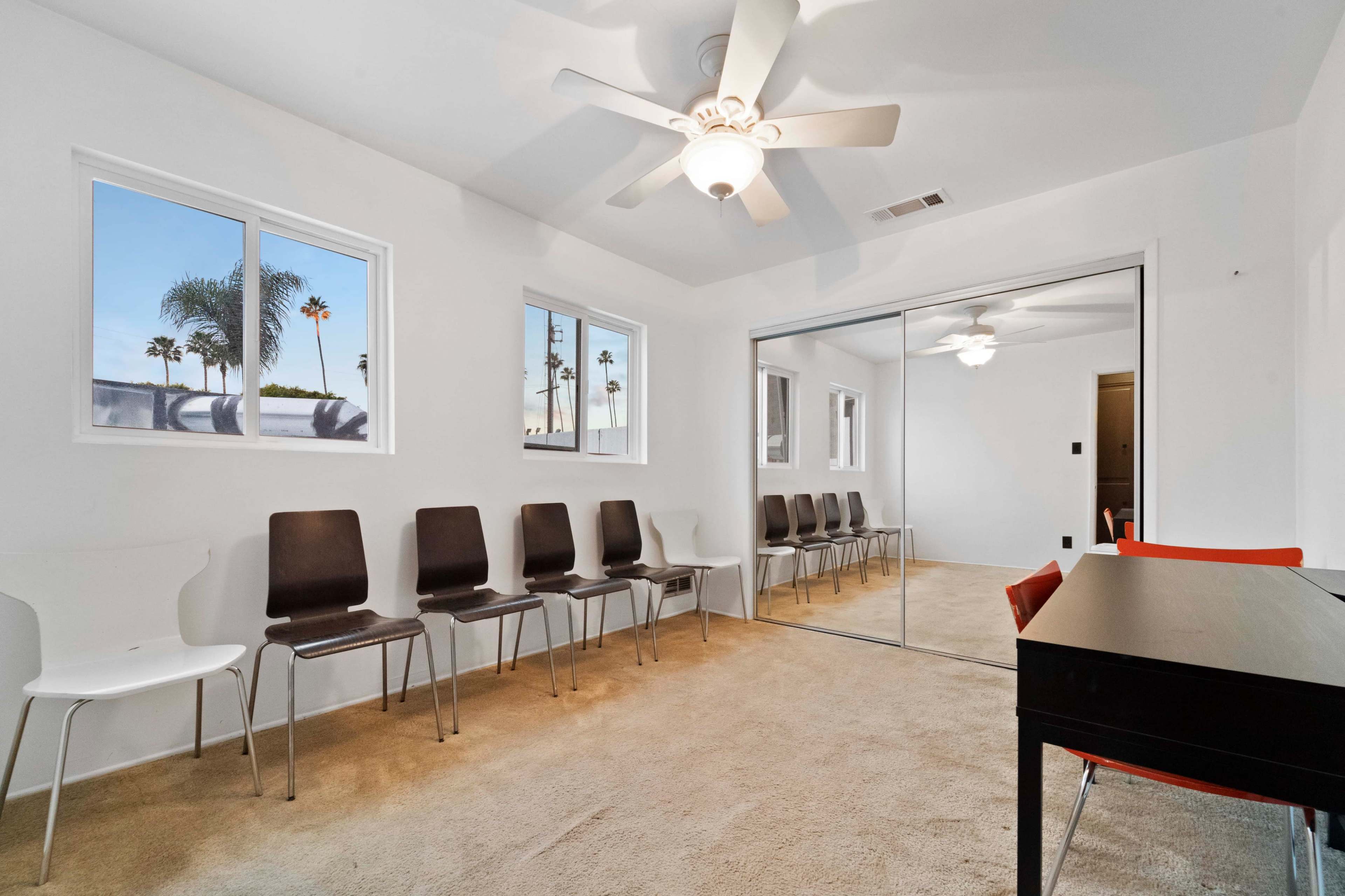 A room with beige carpet, featuring a group of eight chairs against the wall, a desk, and large windows allowing natural light.
