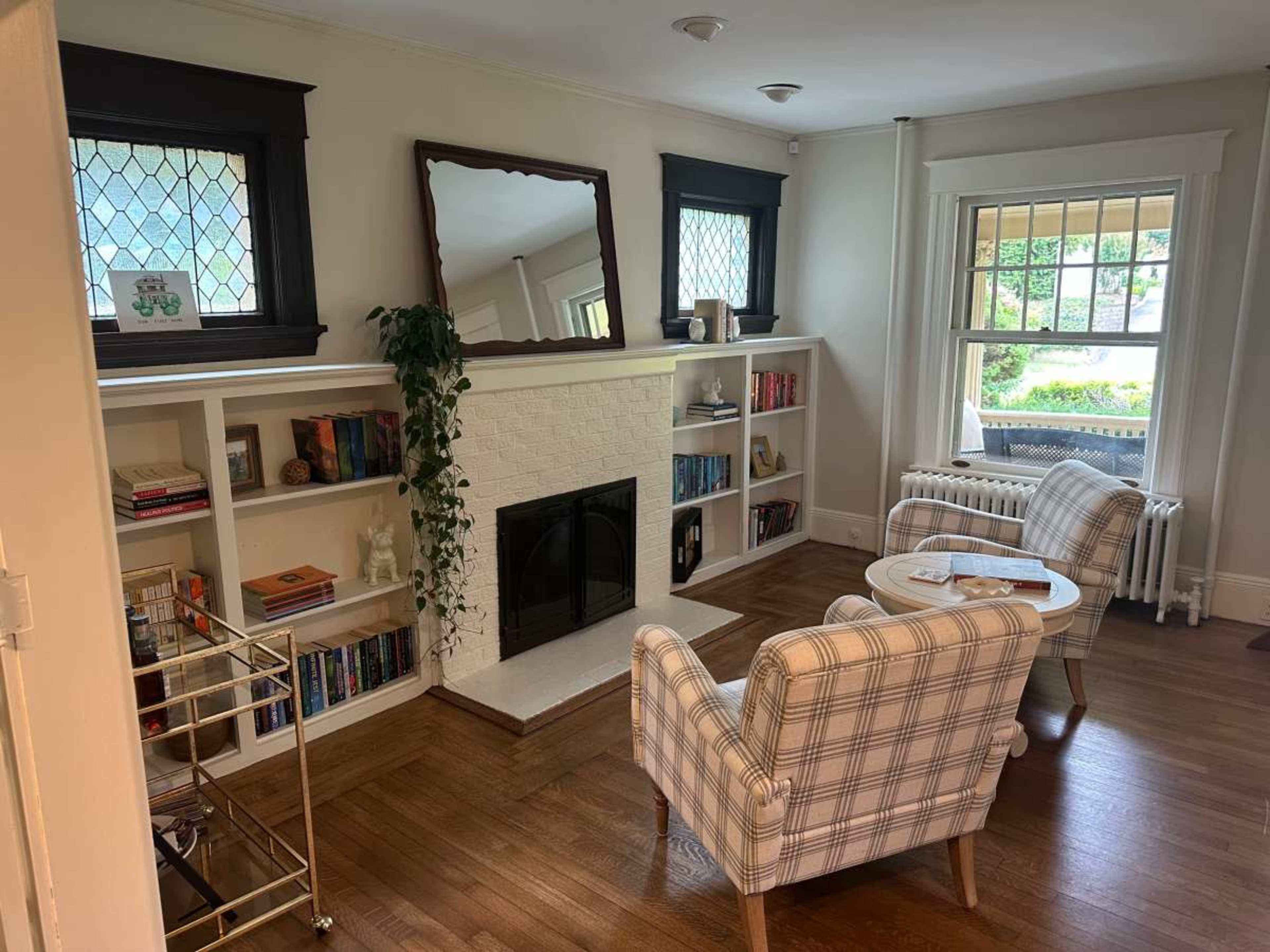The image shows a cozy living room with a fireplace, built-in shelves filled with books, and two armchairs in a plaid pattern.