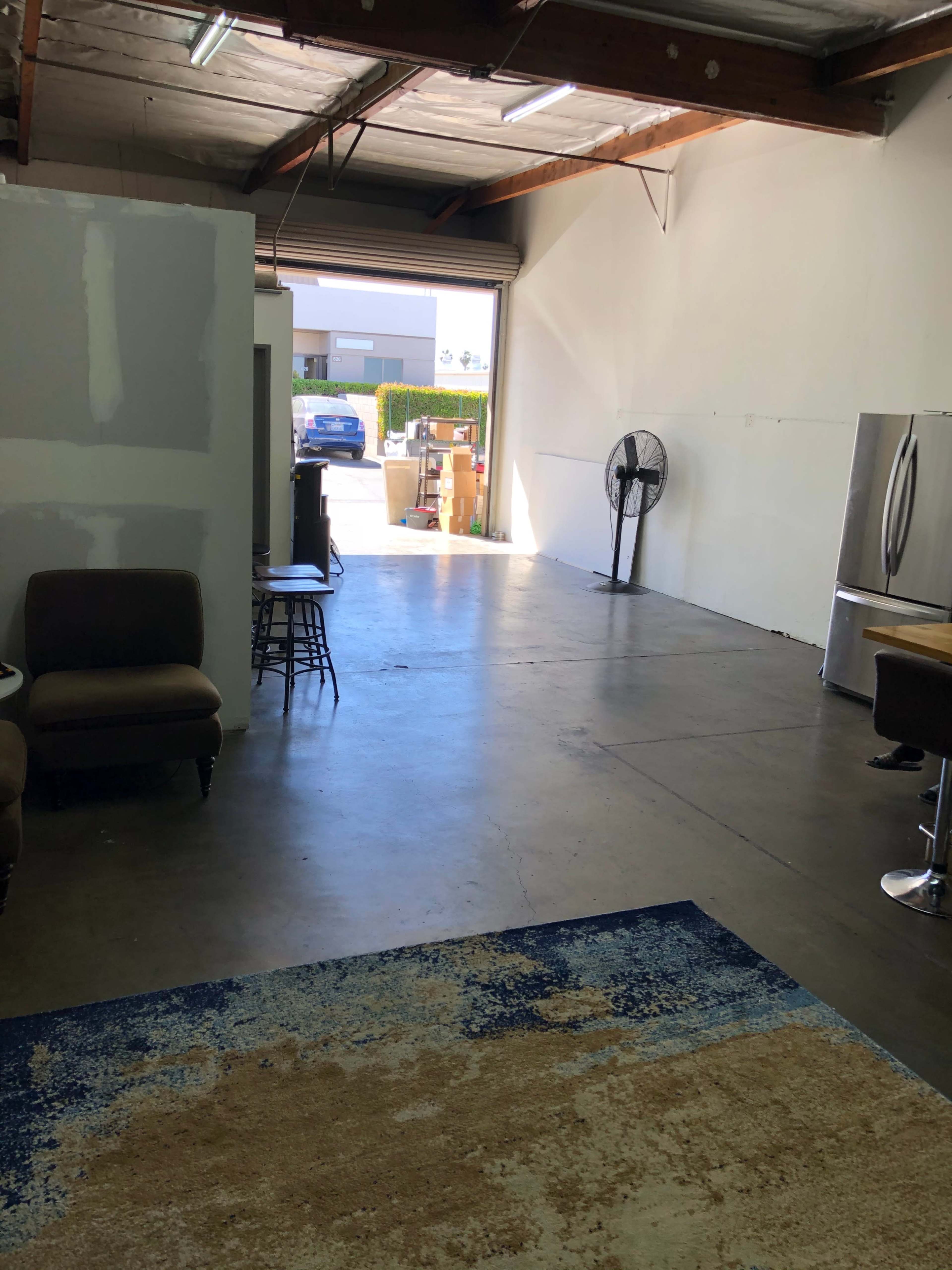 The image shows an empty industrial space with a concrete floor, a large wall fan, a kitchen area with a refrigerator, and a door leading to the outside.