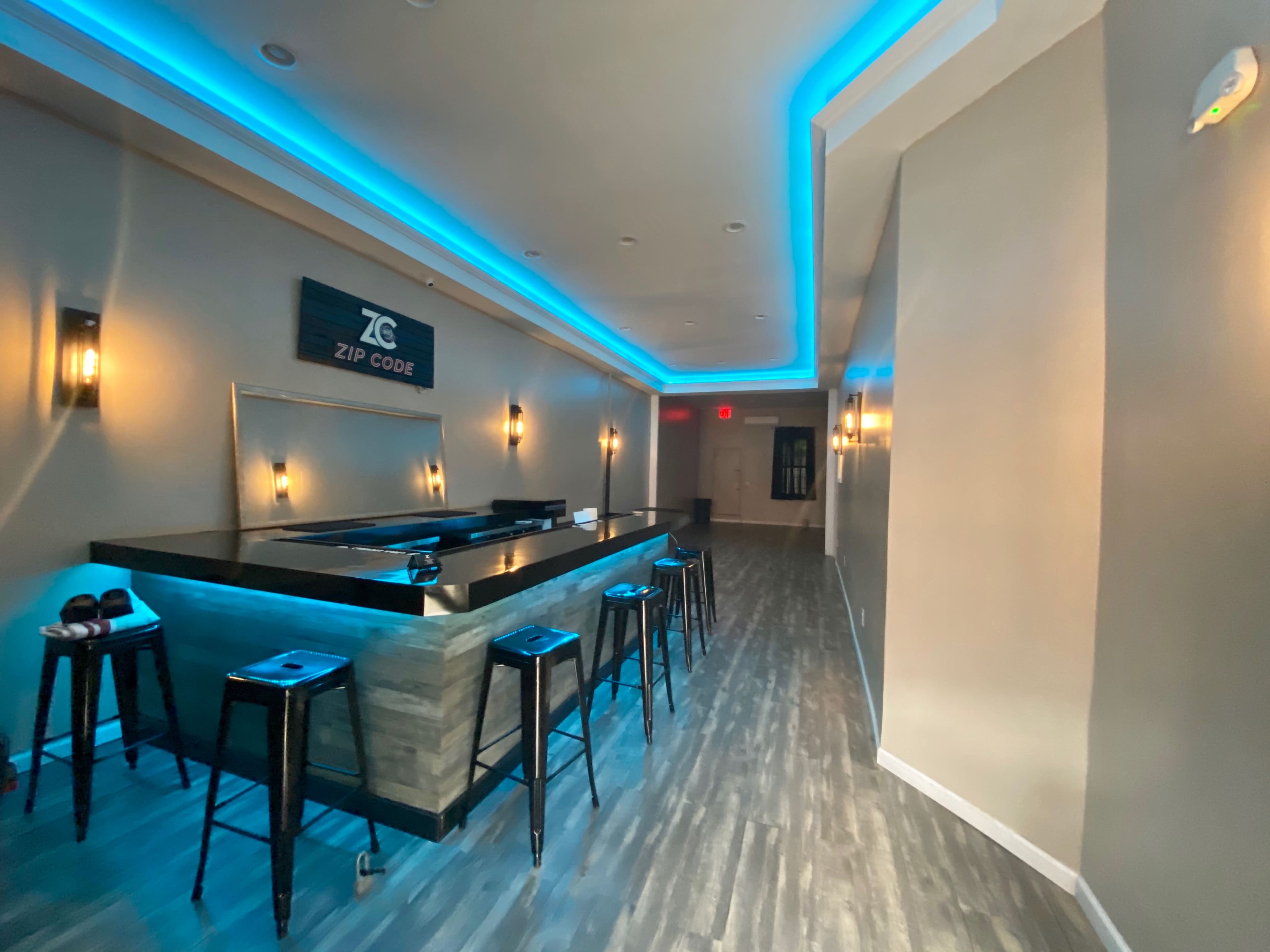 The image shows a modern bar area with a wooden counter and high stools, illuminated by blue LED lights along the ceiling and behind the bar.