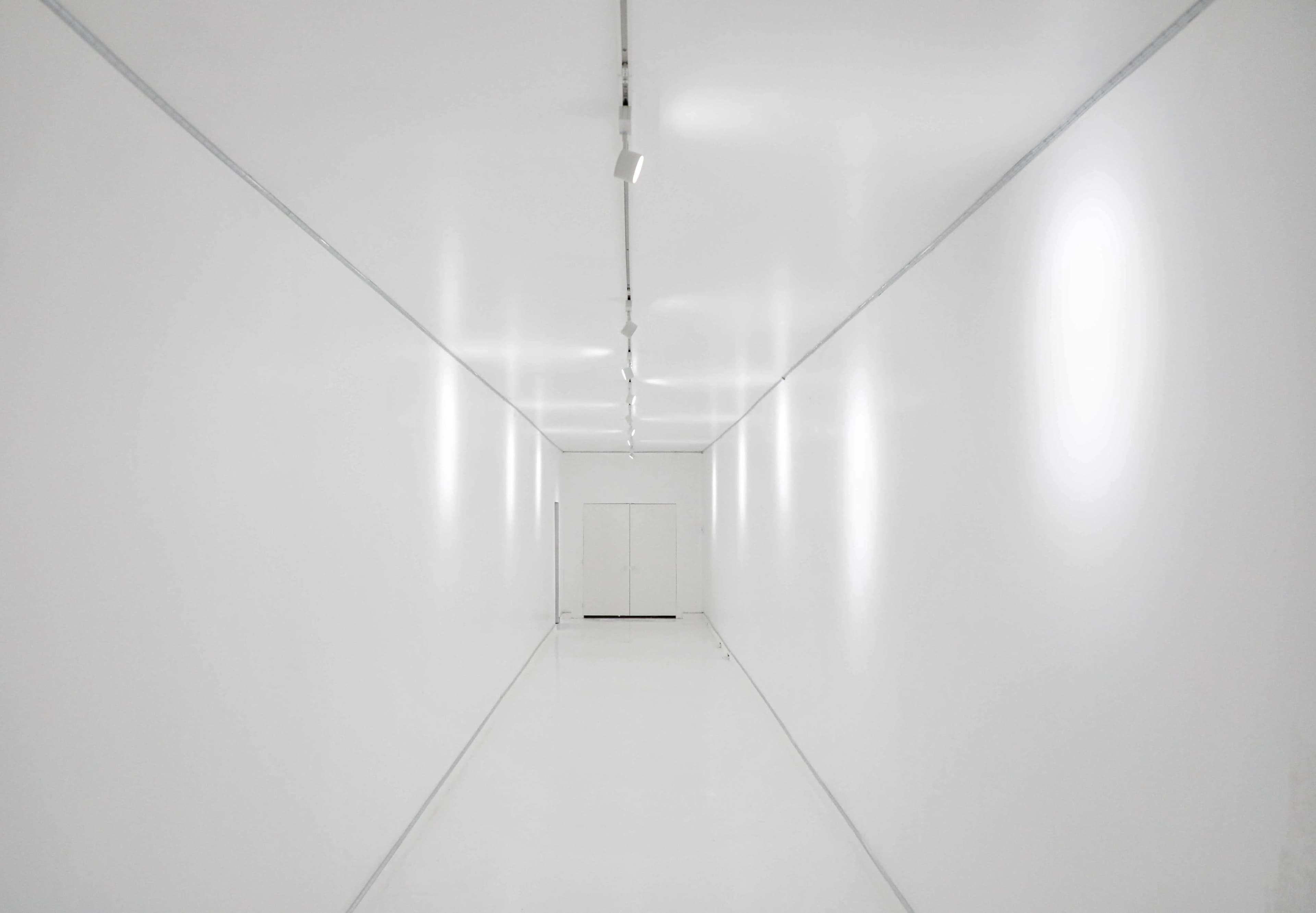 A long, empty hallway features white walls and a glossy white floor, with lights mounted along the ceiling illuminating the space.