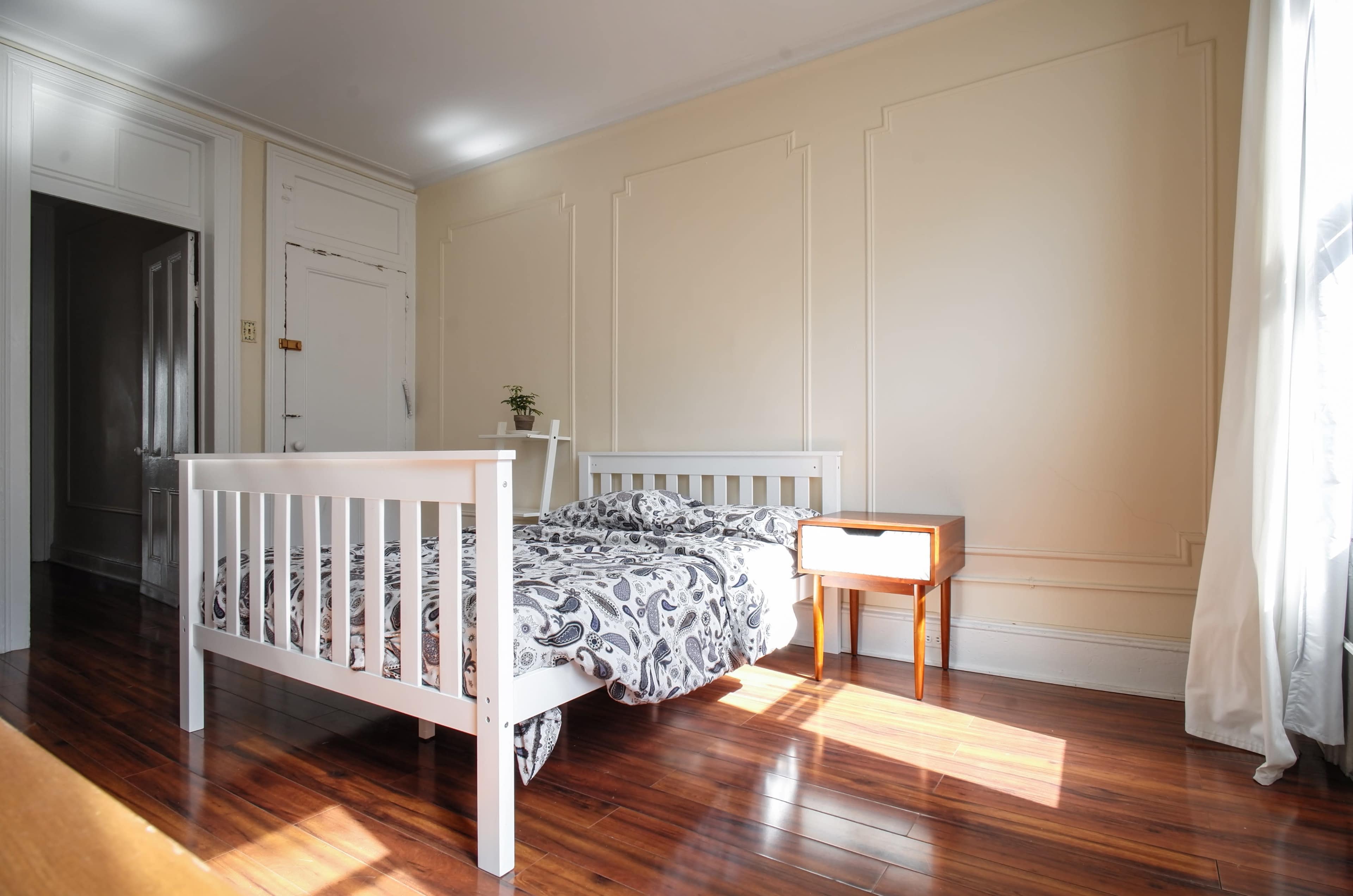 A sunlit bedroom features a white wooden bed with patterned bedding and a small nightstand beside it on polished hardwood floors.