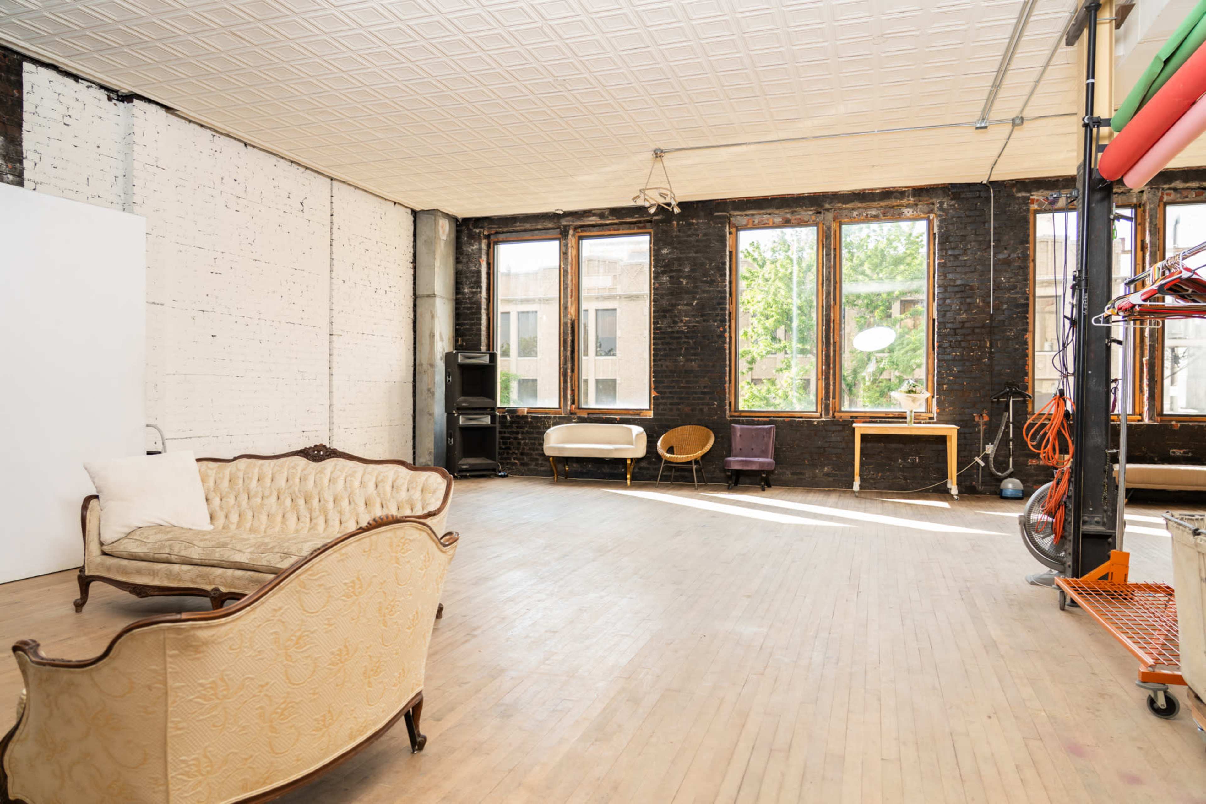 The image shows a spacious room with wooden floors, large windows, and a mix of vintage and contemporary furniture along the walls.