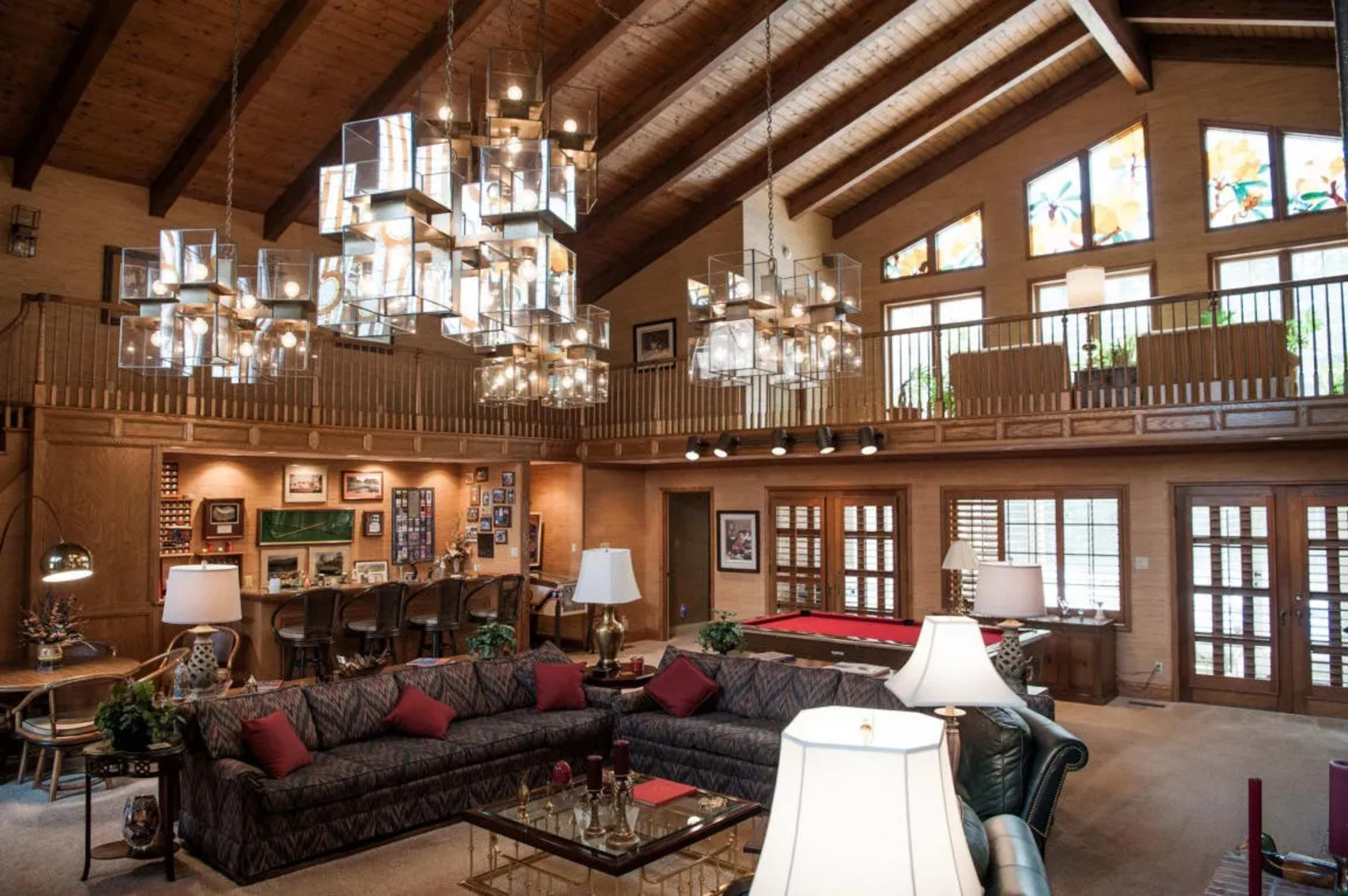 A spacious, well-lit living room with a high wooden ceiling, a large chandelier, and a mix of seating arrangements, including a sectional sofa and a pool table.