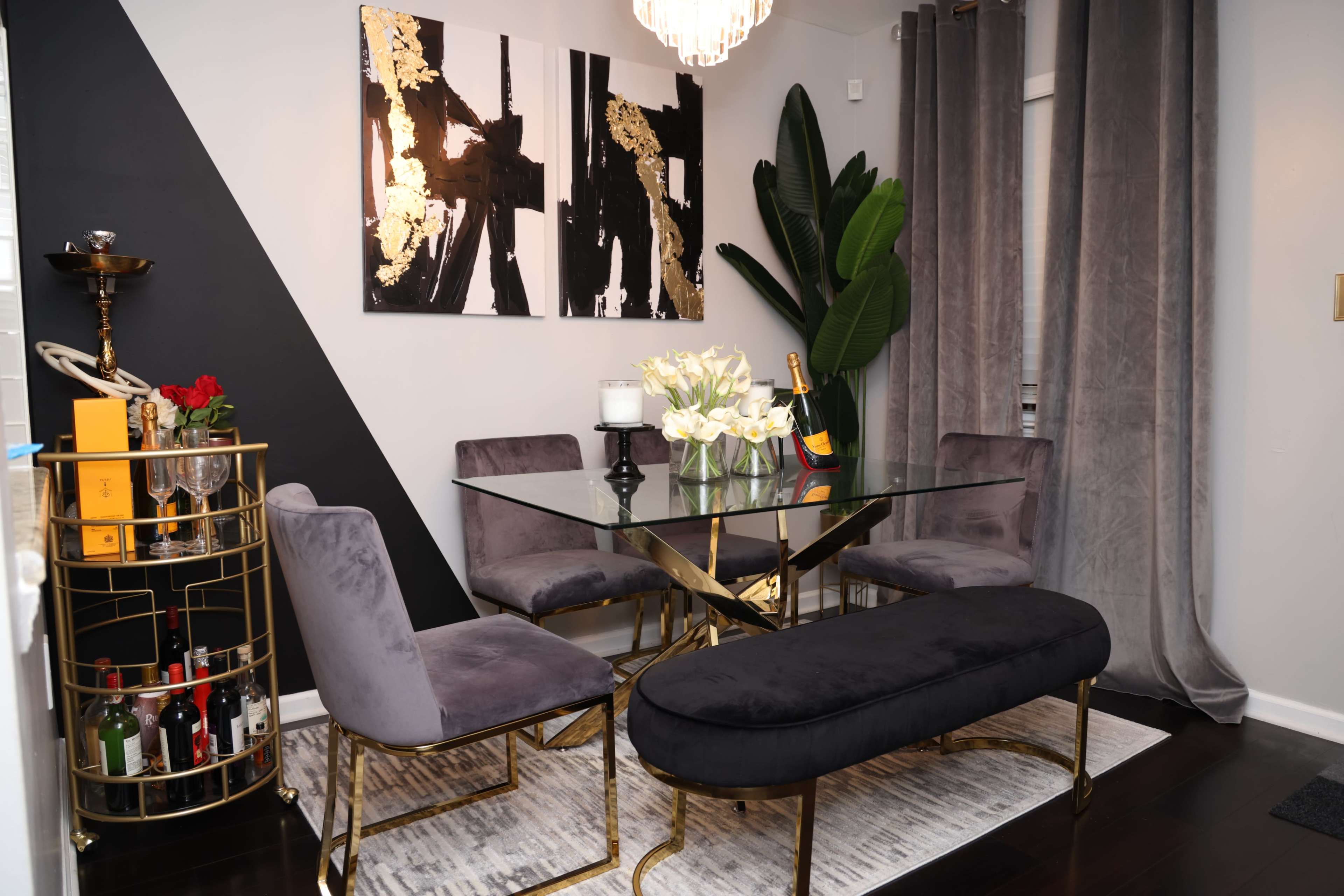 A modern dining area features a glass table surrounded by purple chairs, a gold-framed bar cart, and artwork on the wall alongside a tall plant and grey curtains.