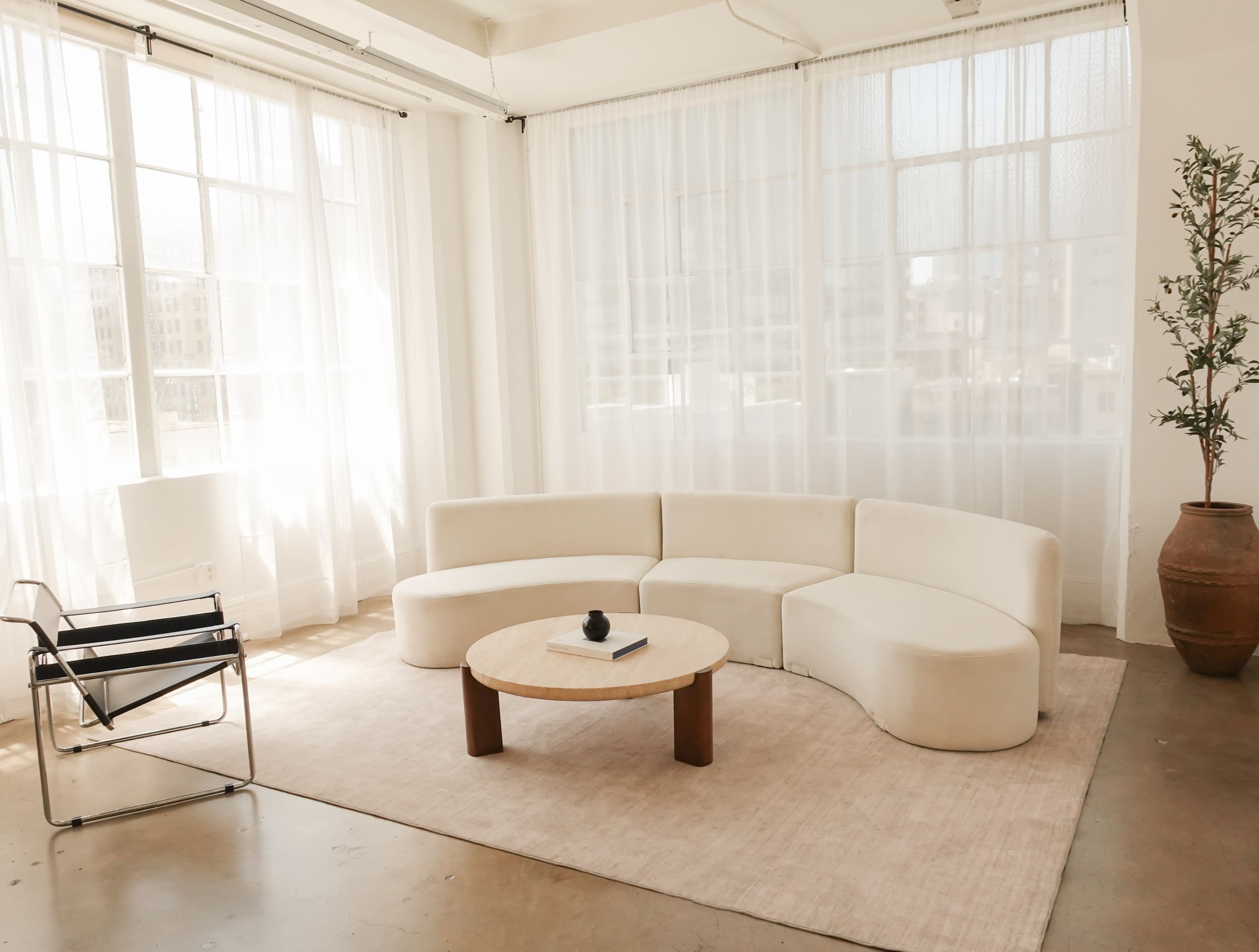 The image features a modern, minimalist living space with a curved white sofa, a round wooden coffee table, and large windows covered by sheer curtains.