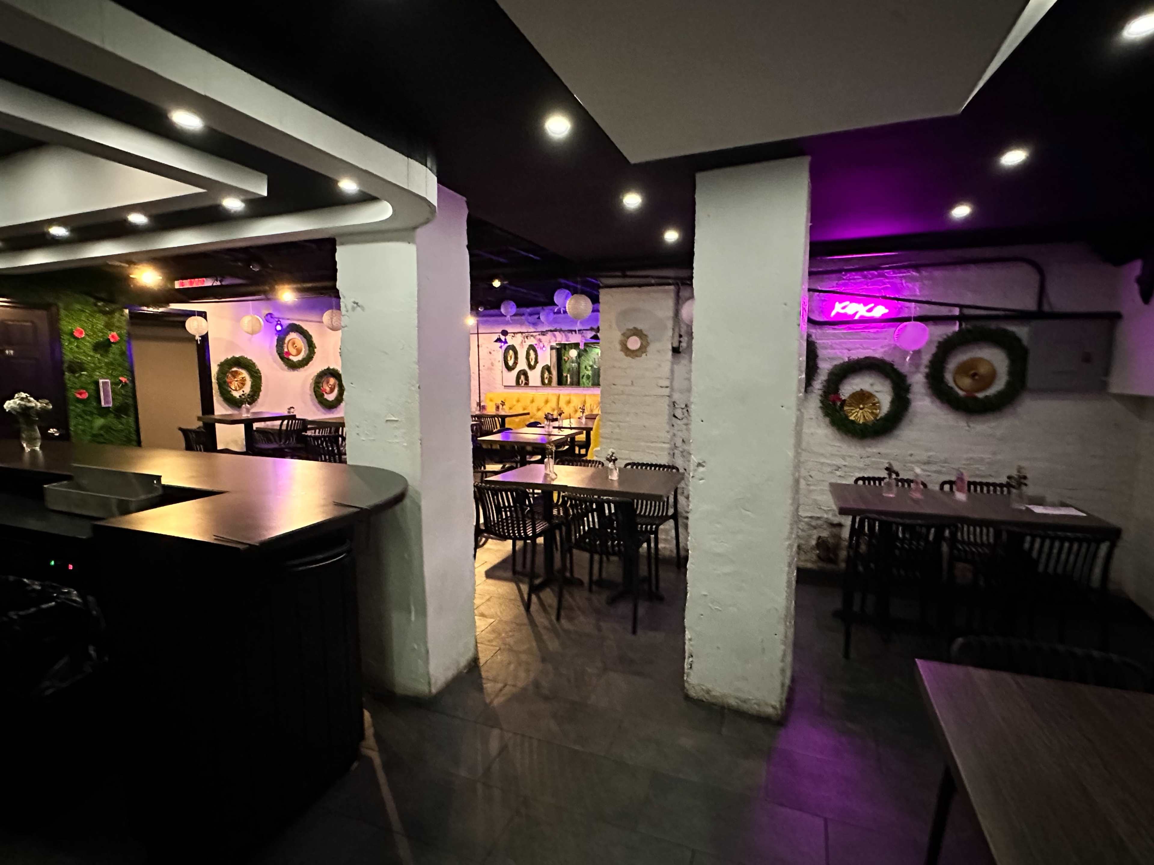A dimly lit restaurant interior with multiple tables arranged in a spacious layout, decorated with circular wreaths and neon lighting.