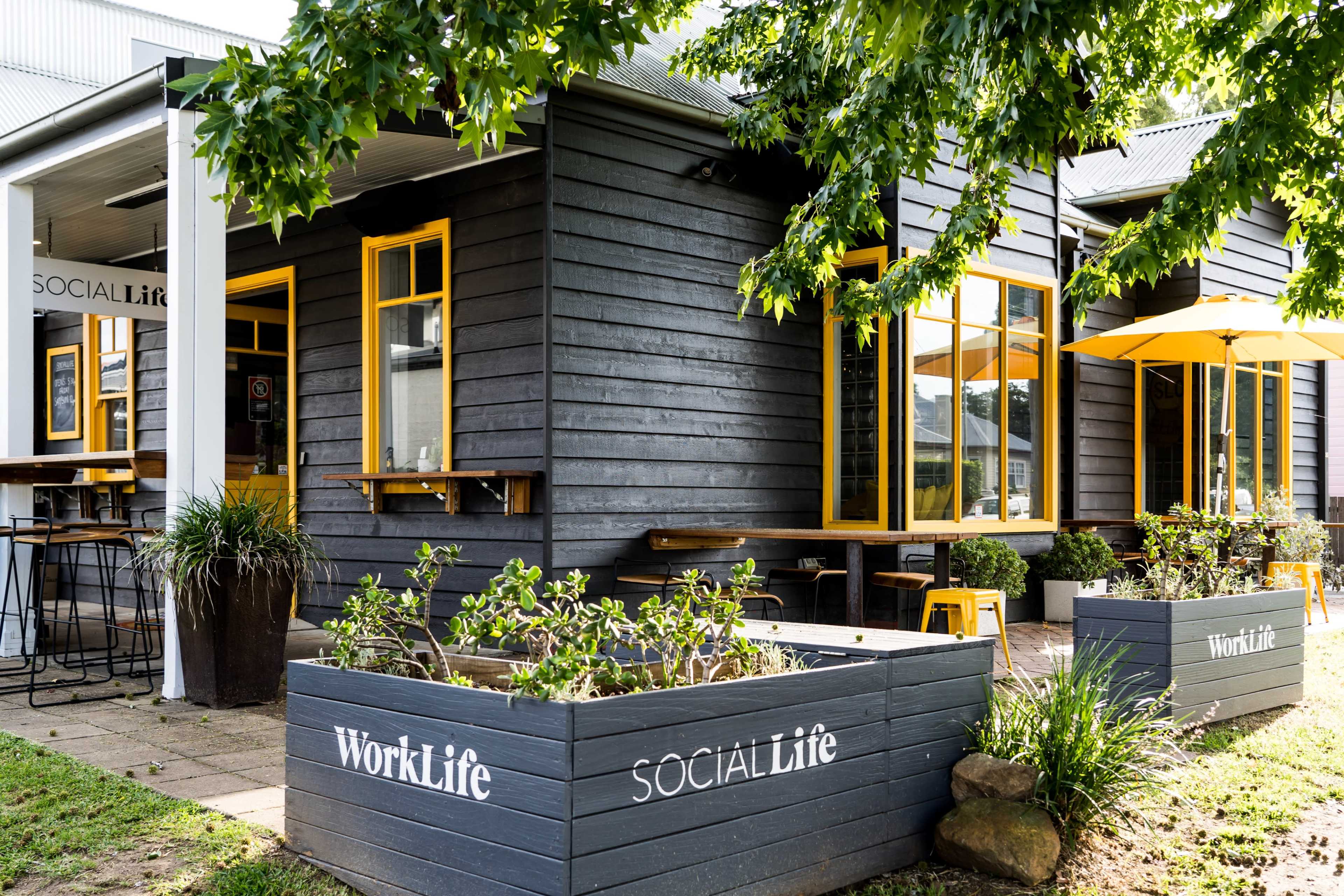 A modern building with dark wooden siding features large yellow-framed windows and signs reading "WorkLife" and "SocialLife," surrounded by greenery and outdoor seating.