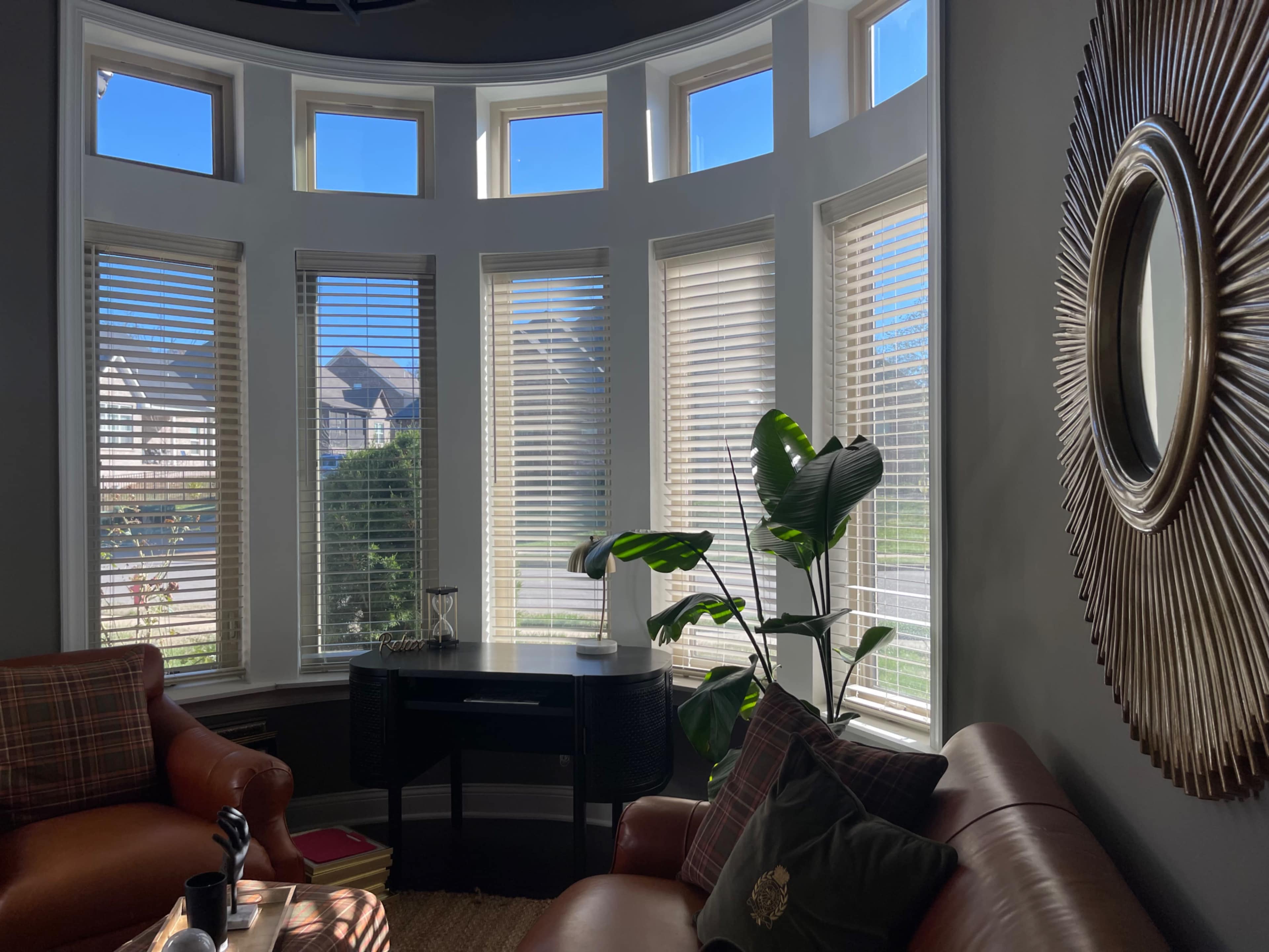A cozy living room features large bay windows with wooden blinds, a brown leather sofa, and a round mirror on the wall.