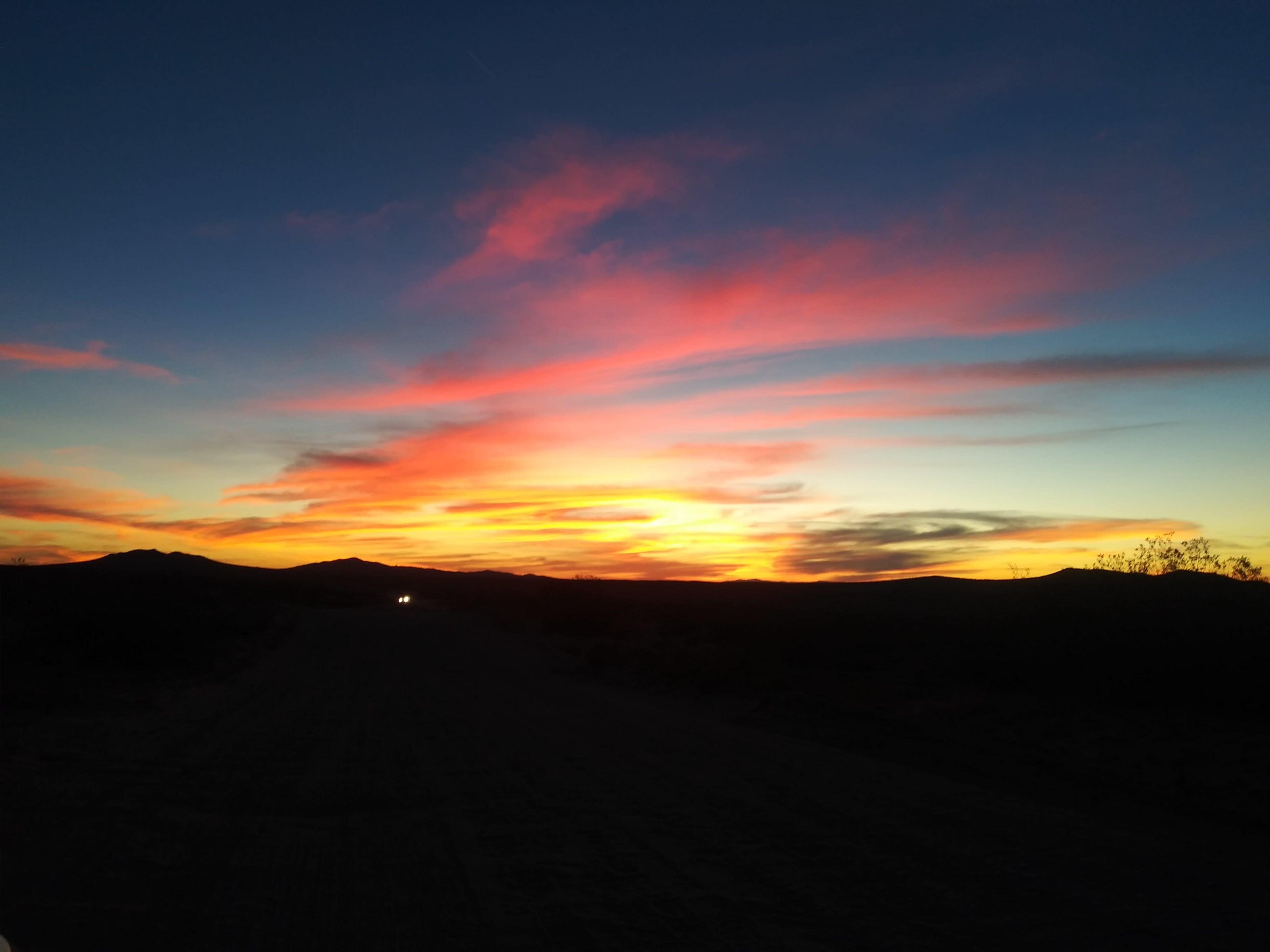 A vibrant sunset casts colorful hues over a desert landscape, with a distant vehicle illuminating the dirt road.